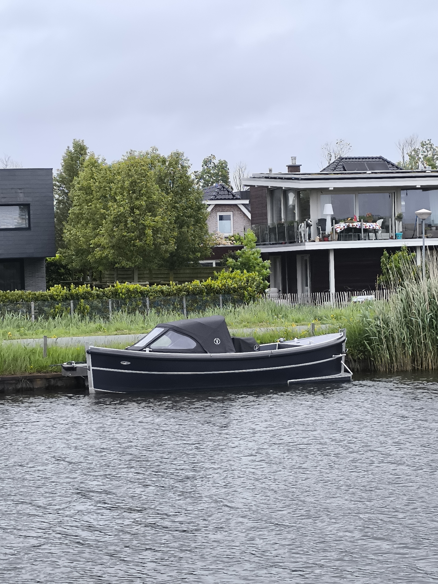A black boat with a black canopy and silver trim is docked on a body of water. The boat is near a grassy shoreline with tall reeds. Behind the shoreline, there are houses and trees. The sky is overcast. The location is Oudorp, Netherlands.