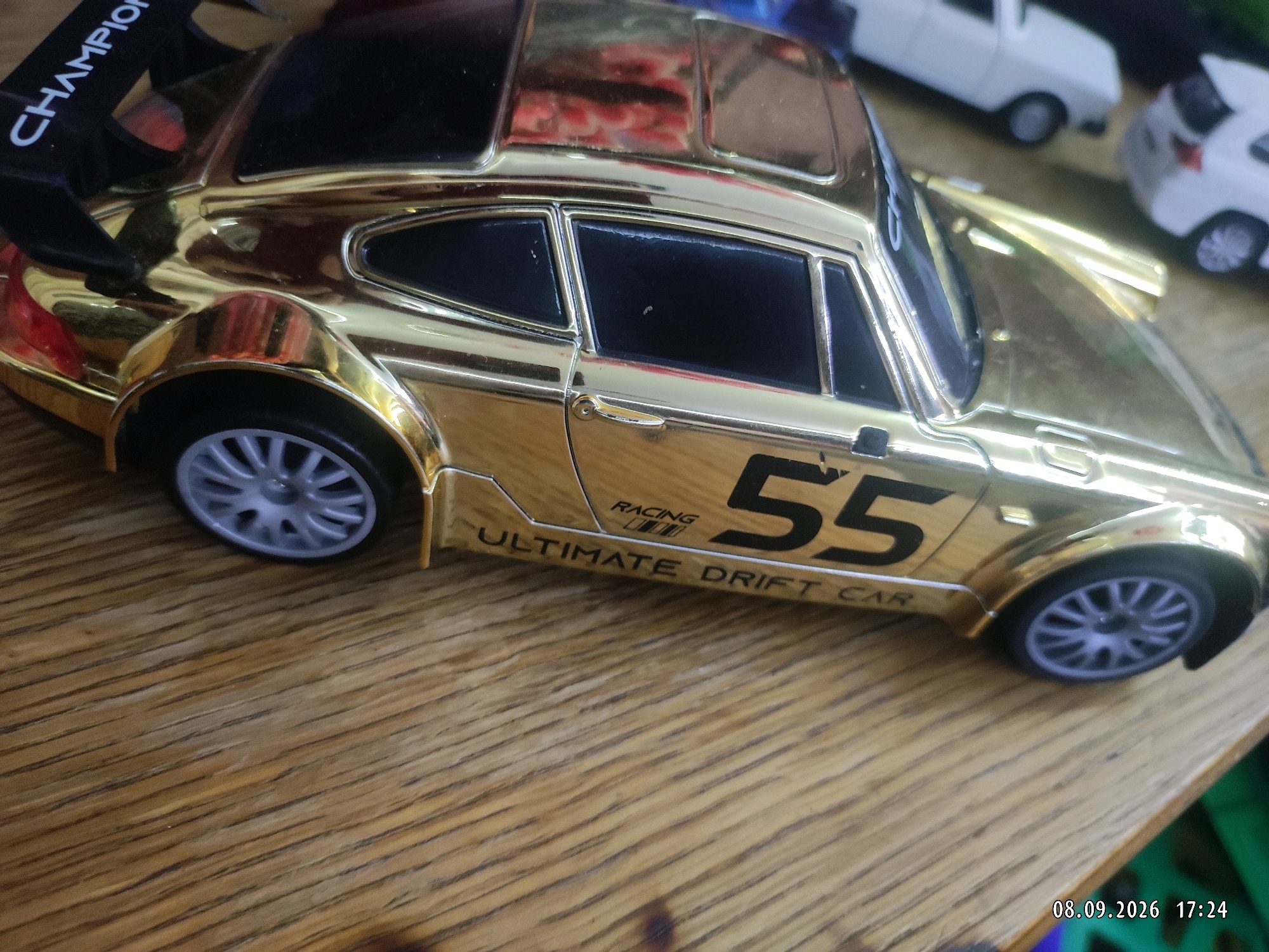 A close-up, high-angle shot shows a gold-colored toy car with a spoiler that reads "CHAMPION" in white text. The car also has the number "55" on its side and text that reads "RACING" and "ULTIMATE DRIFT CAR". The car is on a wooden surface, possibly a table. In the background, out of focus, are other toy cars, one white and one silver. The timestamp "08.09.2026 17:24" is visible in the bottom right corner of the image. There is no indication that this scene is located in Makhachkala, Russia.