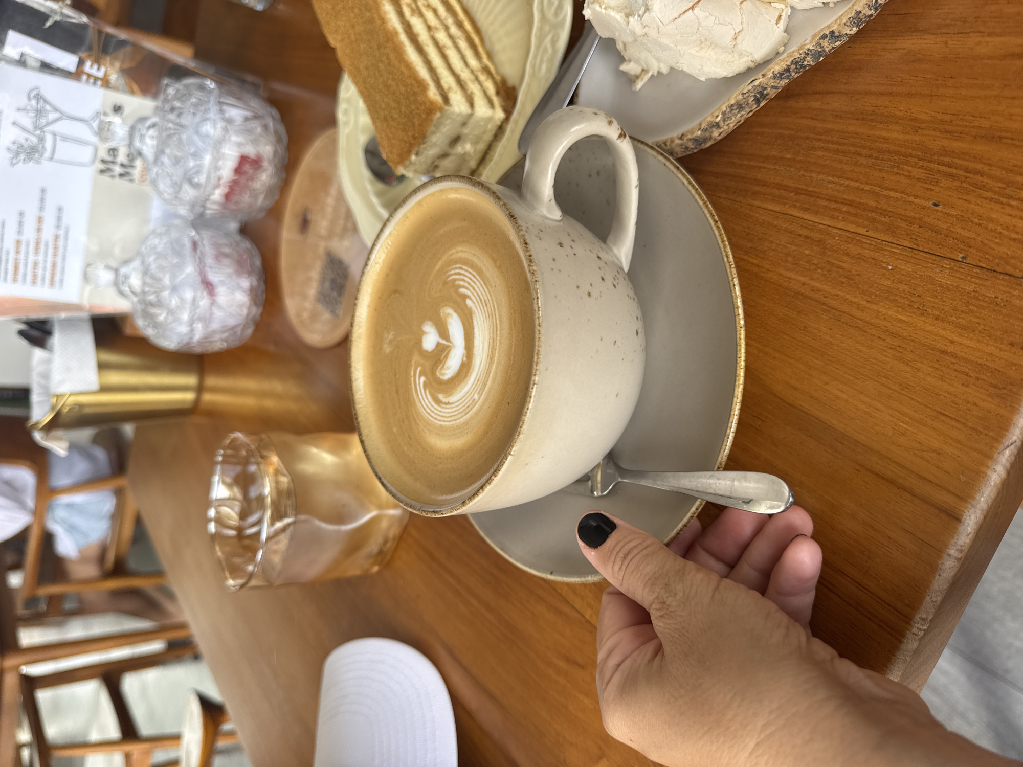 A hand with black painted fingernails is reaching for a spoon resting on a saucer in front of a cup of coffee with latte art. The coffee cup is speckled beige and sits on a gray saucer. To the right of the coffee is a slice of layered cake on a yellow plate, and next to that, a white dessert on a rough-edged plate. A glass of amber liquid sits behind the coffee cup. In the background, two ornate glass jars with metal lids are visible, as well as a gold bucket. The scene is set on a wooden table, likely in a cafe in Kuta Selatan, Indonesia. The focus is on the coffee and the hand, with the background elements slightly blurred.