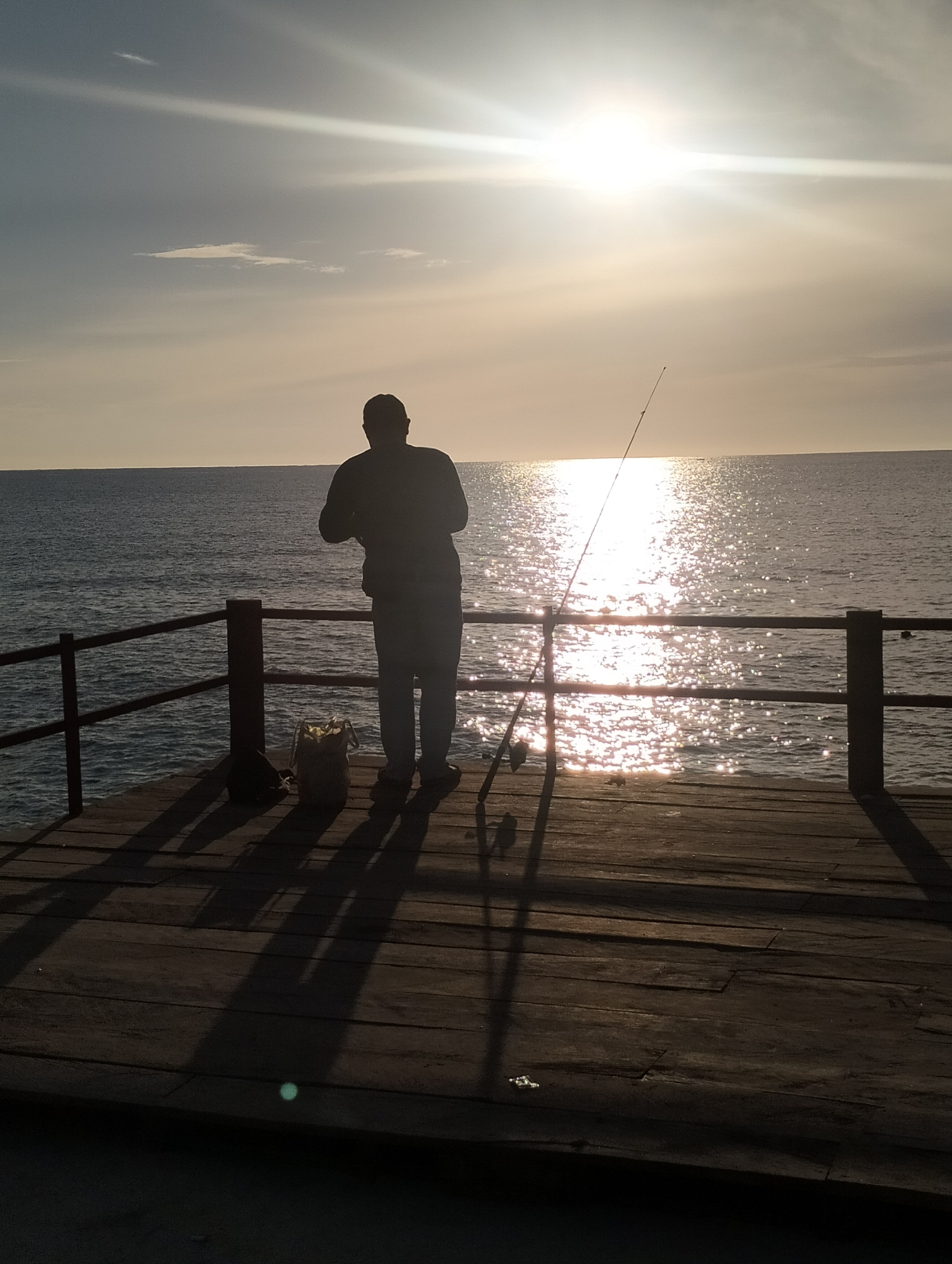 A lone fisherman stands on a wooden pier, silhouetted against a bright, hazy sun setting over the ocean. The sun's rays create a glittering reflection on the water's surface. The fisherman, facing away from the camera, holds a fishing rod, casting a long shadow onto the pier. A fishing bag is placed on the pier near him. The pier has a wooden railing. The scene appears to be at a coastal area in Lhokseumawe, Indonesia, given the tropical feel and the body of water.