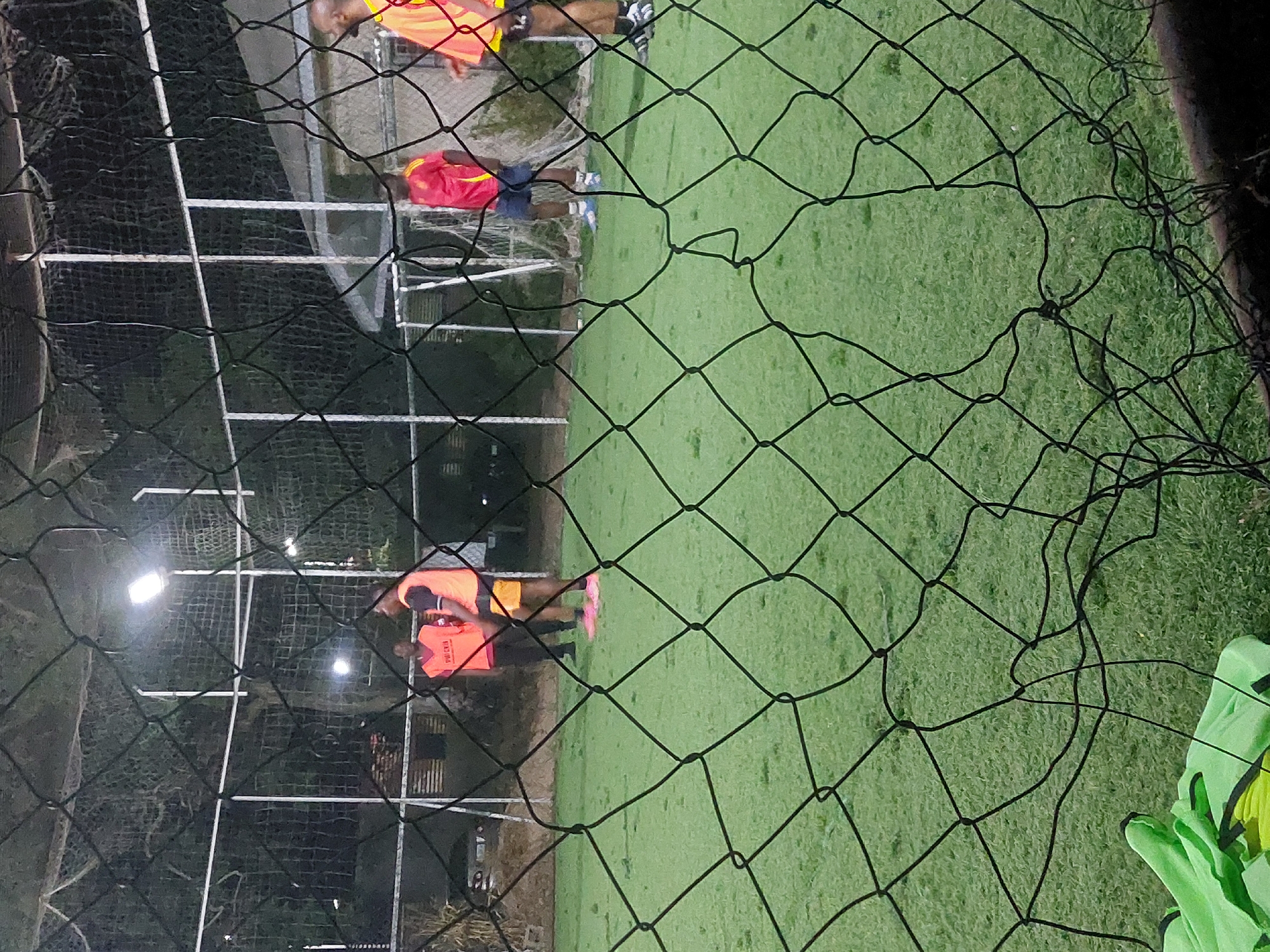 The image depicts an outdoor artificial turf sports field at night, observed through a black net in the foreground, in Bar Jirgi Summa, Nigeria. The field is illuminated by several bright overhead floodlights, visible in the background. Four adult males are present on the green turf.
From left to right:
1. A male figure wears an orange vest with "PUCKY" or similar text, over a dark shirt and dark trousers, standing facing the right. His footwear is not clearly discernible.
2. A second male figure wears an orange sleeveless top and dark yellow shorts, with pink athletic shoes. He stands facing left, with his back slightly turned to the camera.
3. A third male figure, positioned further back and to the right, wears a red short-sleeved shirt with yellow stripes on the shoulders, blue shorts, and blue and white athletic shoes. He faces left.
4. A fourth male figure, partially visible on the far right, wears an orange sleeveless top with yellow trim, dark shorts, and black and white athletic shoes. He is positioned facing left.
All individuals appear to be standing still; no active play is visible. In the bottom left foreground, a folded light green or yellow fabric item is resting on the artificial turf, close to the foreground net. Beyond the field, structures, trees, and additional fencing or netting are visible in the dim background light.