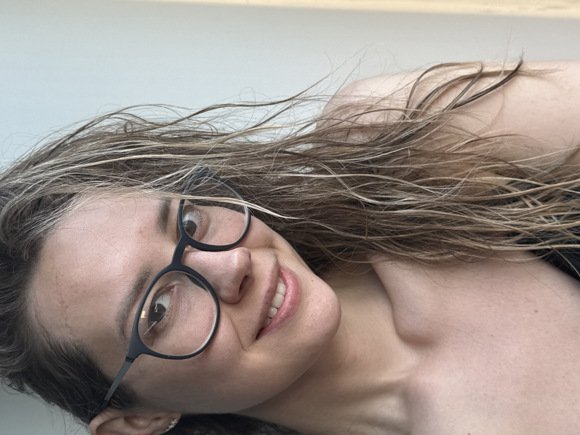 A fair-skinned female wearing black-rimmed glasses is smiling at the camera. Her hair is wet and blonde with darker roots. The background is a plain off-white color, and the subject's shoulder is visible. There is no information to indicate the location is in Chaikovsky, Russia.