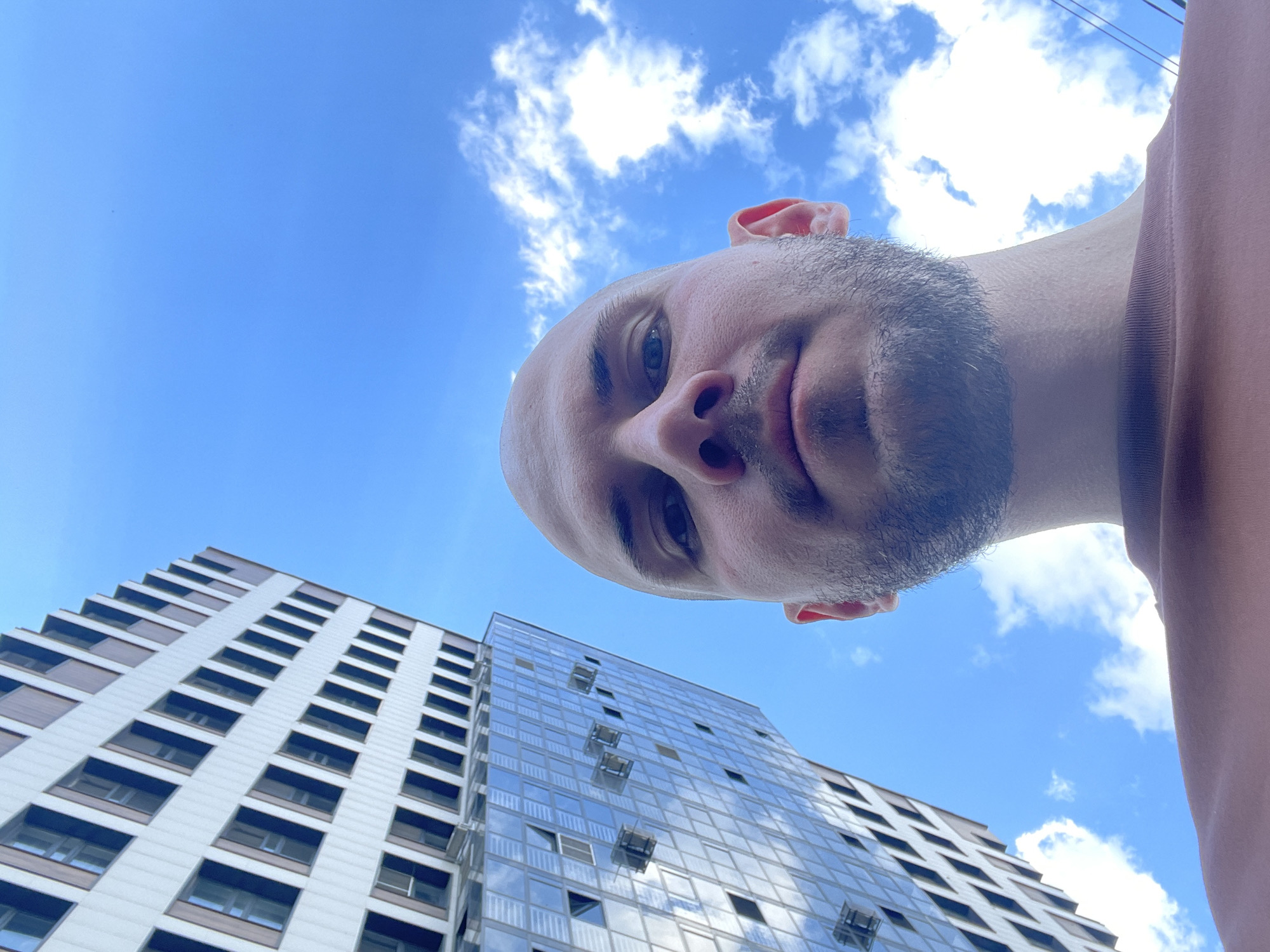 A man is looking down at the camera from a low angle, with two tall buildings visible in the background against a blue sky with scattered clouds. One building is predominantly white with dark horizontal accents under each window; the other has a glass facade. Based on the architecture and context clues (the man's apparel and coloring), this is likely in Krasnoyarsk, Russia. There are power lines in the top right of the image. The man is bald with a short beard, and is wearing a brown shirt.
