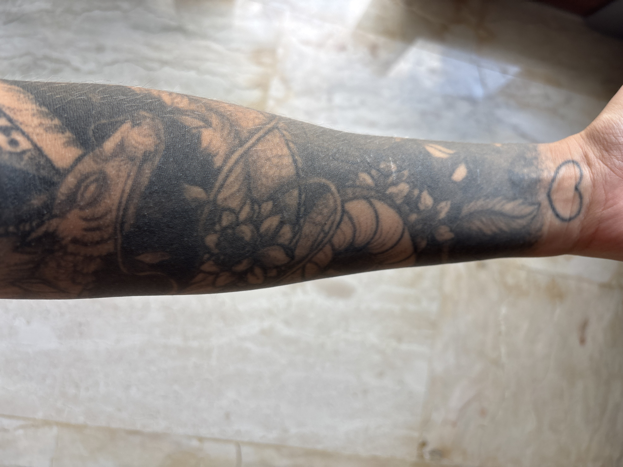 An arm, heavily tattooed with black ink, stretches across the frame. The tattoos include floral elements and abstract shapes. On the wrist, a small, simple heart outline is visible. The arm rests on a surface composed of light-colored, possibly marble, tiles.
The location of the photograph is unknown, and there is no reference to Cemagi, Indonesia.