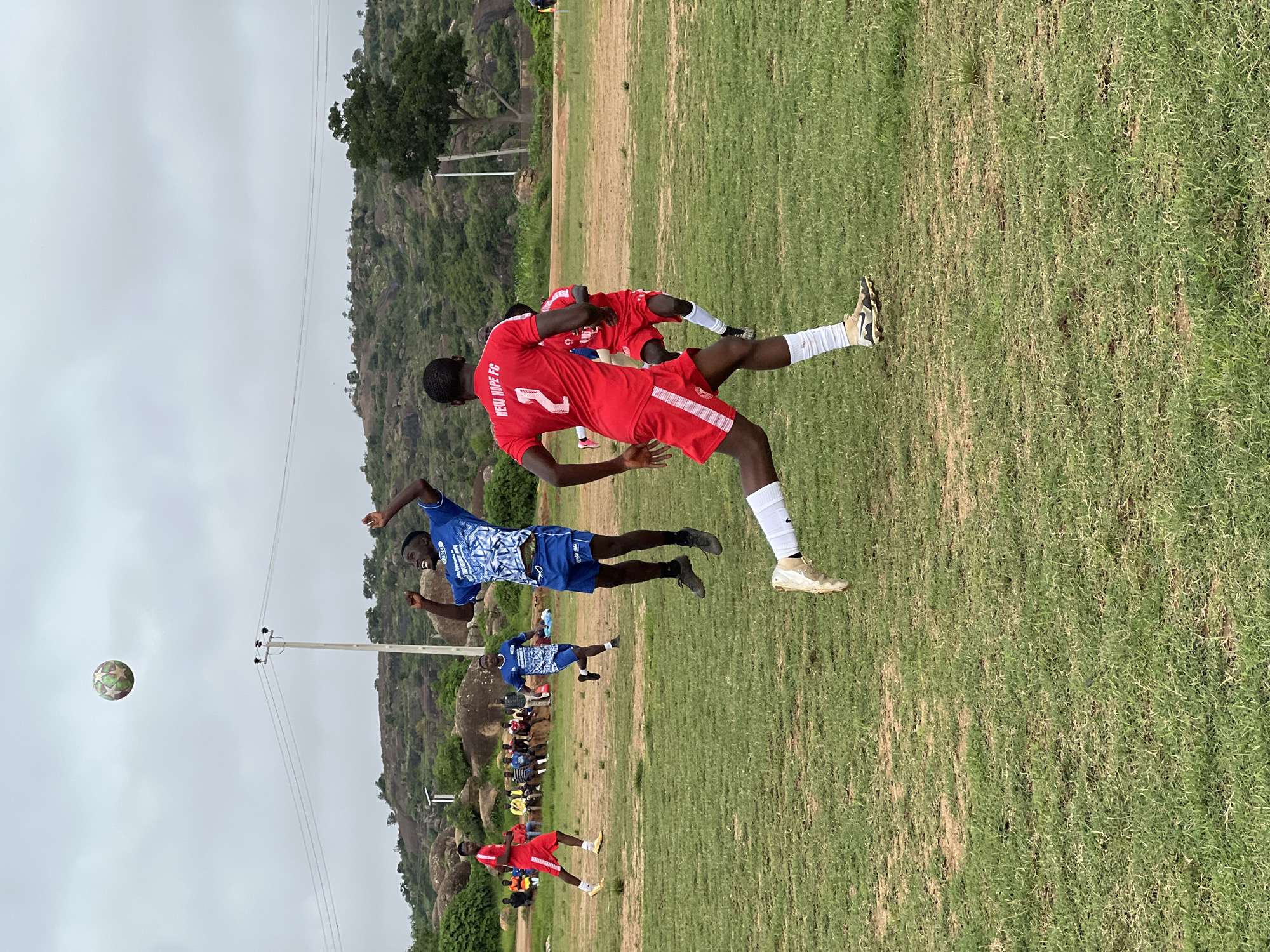 A group of young men, some in soccer uniforms, are playing soccer on a grassy field. The field is bordered by rocky, hilly terrain with scattered trees and vegetation. In the background, there are spectators sitting on benches, and a utility pole with wires is visible. The sky is overcast. The players are engaged in active play, with one player in a blue jersey mid-air reaching for a soccer ball. Another player in a red jersey with the number "2" and "NEW HOPE FC" on the back is running. The scene appears to be a soccer match taking place outdoors in a somewhat undeveloped area, likely a community field, consistent with the landscape often found on the outskirts of Jos, Nigeria.