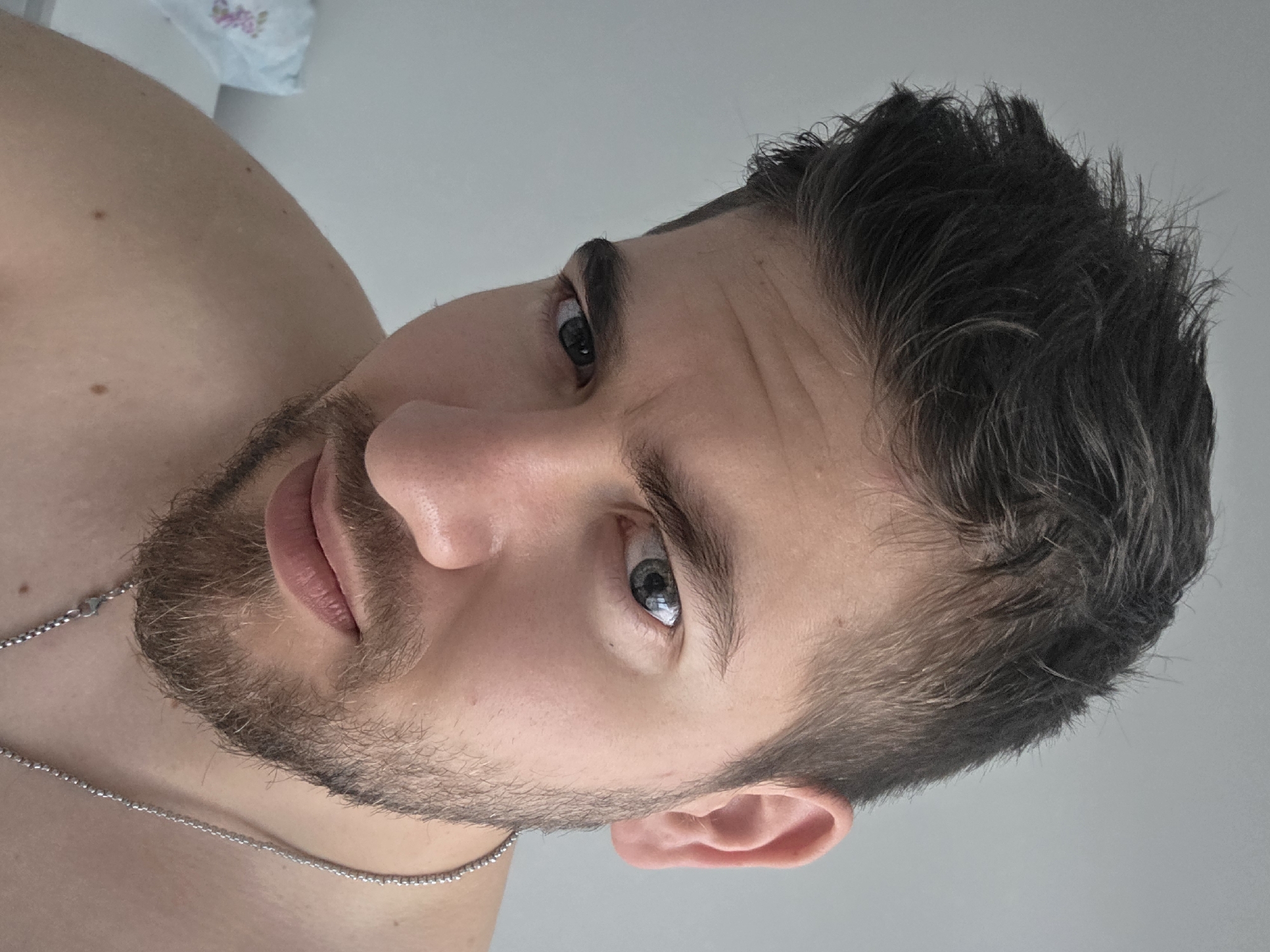 A close-up, angled shot of a man's face and upper chest. The man has dark hair and a beard, and his eyes are light-colored. He is wearing a thin silver chain around his neck. The background is a plain white wall. No specific location in Yagodovo, Bulgaria is discernible from the image.