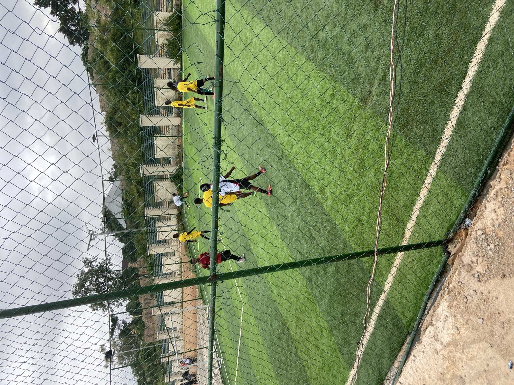 The image shows a group of young people playing soccer on an artificial turf field, likely in Jos, Nigeria. The field is green with white markings, and a chain-link fence surrounds it. In the background, there are buildings, trees, and a rocky hill, consistent with the landscape of Jos. The sky is overcast, suggesting a cloudy day. The players are wearing various colored uniforms, including yellow and red, and some are actively engaged in the game, with one player in a white and black jersey in the foreground, appearing to be dribbling a ball. The overall scene depicts a casual soccer match taking place in an urban or semi-urban environment in Jos.