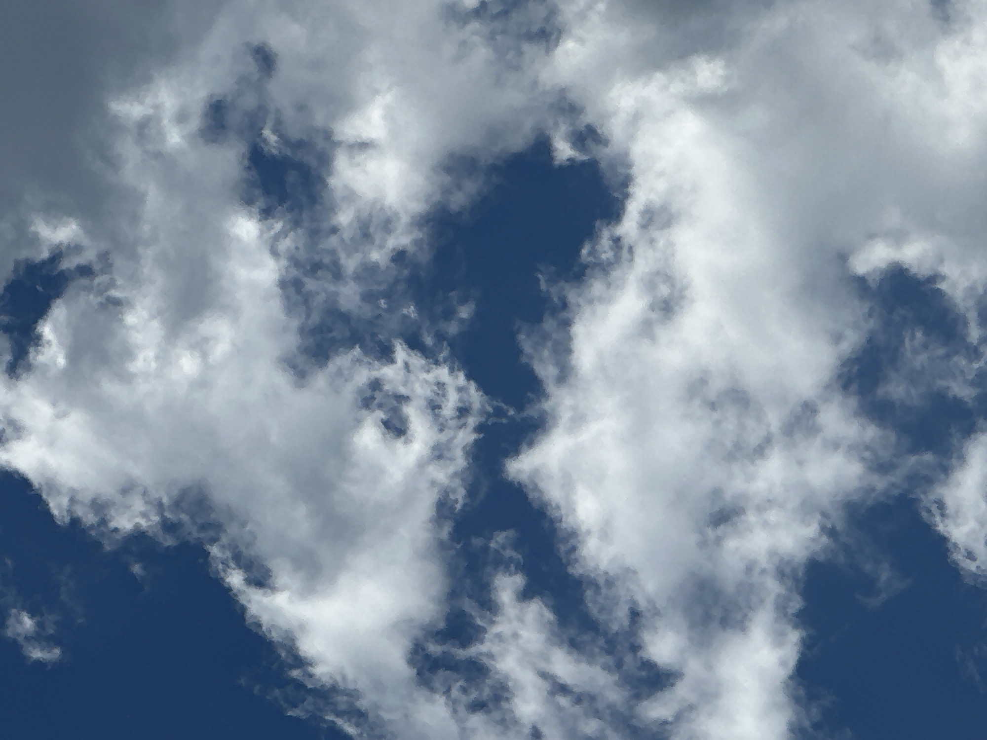 The image depicts a daytime sky filled with scattered clouds. The background is a deep blue, visible in irregular patches between the cloud formations. The clouds are primarily white and various shades of light to medium grey, exhibiting diverse, amorphous shapes and densities. Some cloud sections appear brightly illuminated and dense, while others are more diffuse, wispy, or show darker undersides indicating internal shadowing. No discernible landmasses, structures, vegetation, water bodies, people, animals, or man-made objects are present within the frame. The scene offers no specific geographical or architectural context, rendering the location in the city of Unknown, Unknown, undeterminable from the image content.