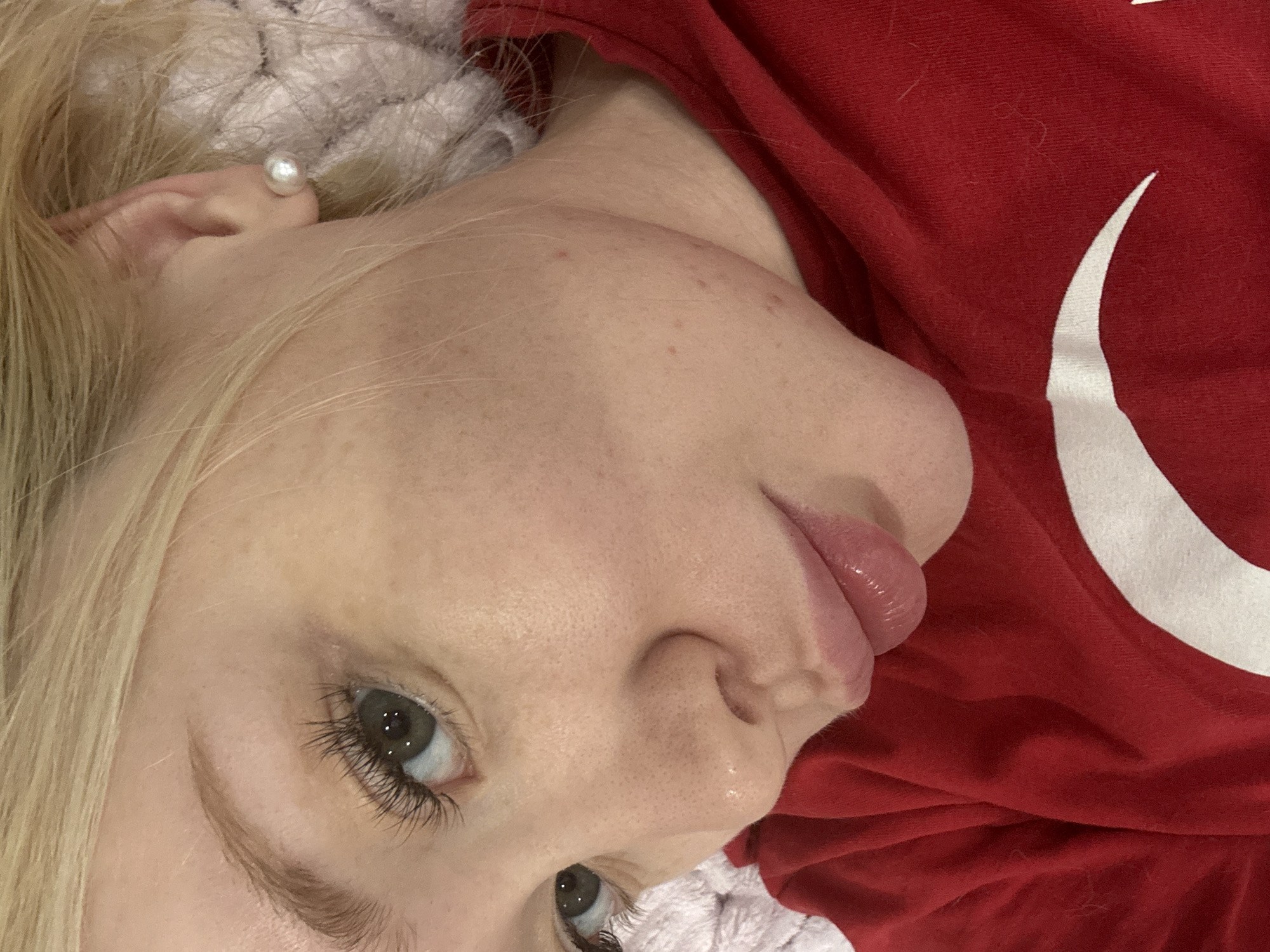 A close-up, overhead, indoor shot shows a young blonde woman wearing a red t-shirt. She is lying down on a pink and white blanket. Her left eye, nose, and mouth are visible. She has light skin, blonde hair, and green eyes. She is wearing a pearl stud earring in her left ear. The t-shirt has a white graphic design on it.