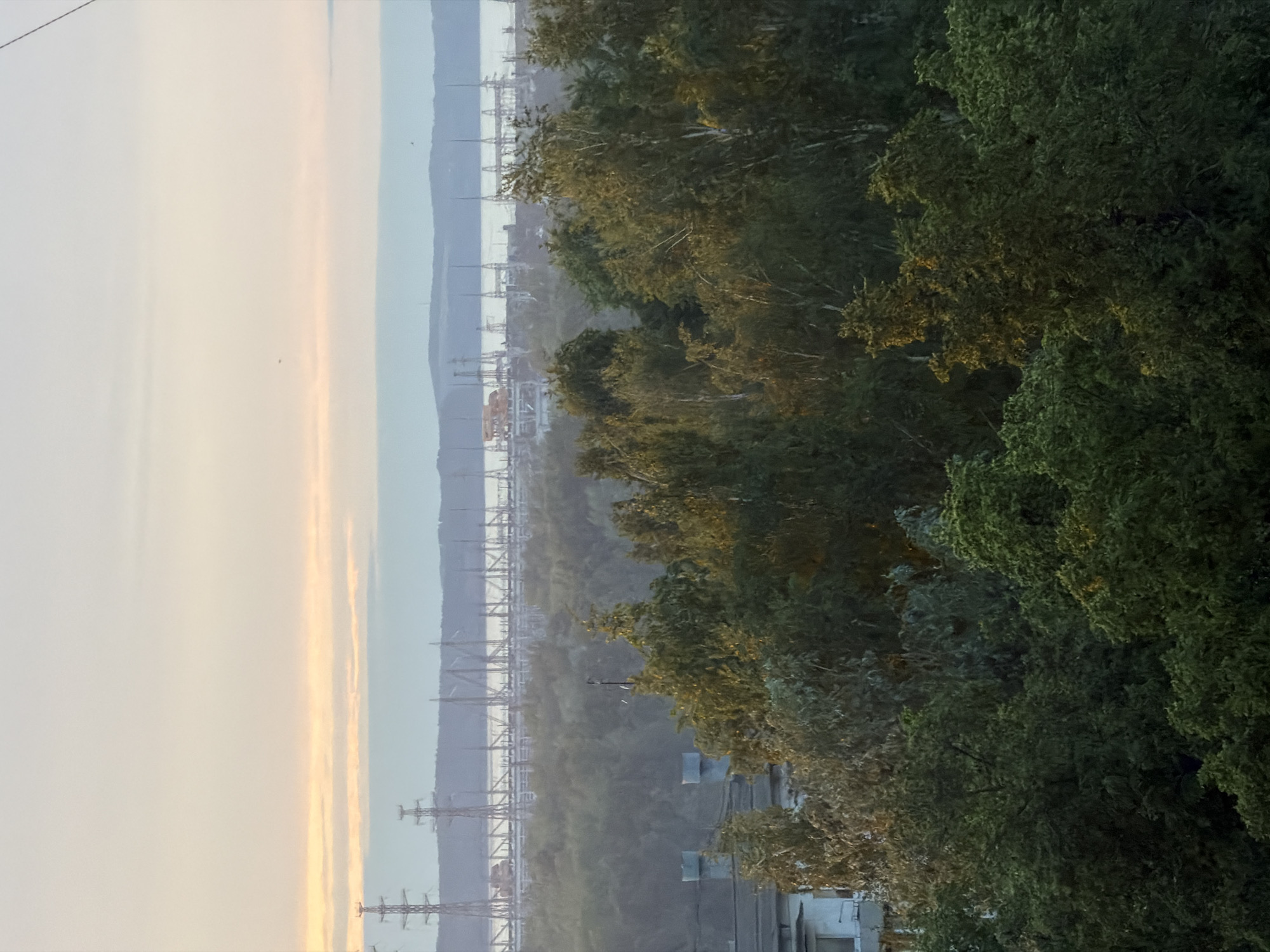 The image shows a landscape in Chaikovsky, Russia, featuring the Kama River. Power lines traverse the foreground and extend across the river towards the distant bank. On the right, dense trees partially obscure a building. The sky is hazy and pale, indicative of early morning or late evening light. No people are visible in the image.