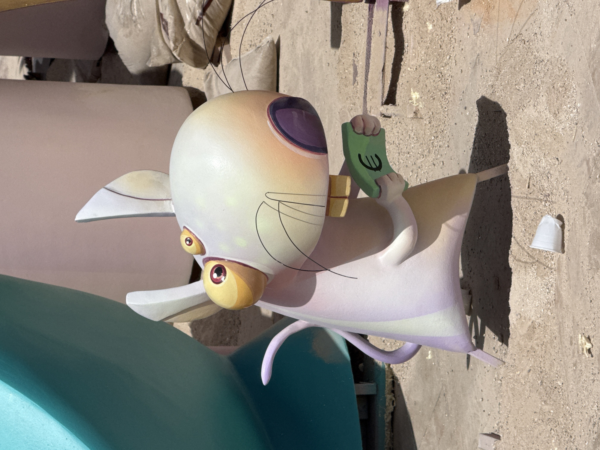 A cartoonish, overweight mouse figure is the main subject of the image. It is depicted with large, round yellow eyes with red pupils, a large white head with pink and yellow shading, and small yellow front teeth. The mouse has large, pointed ears and a thin, purple tail curled behind its back. Its body is predominantly white with faint vertical stripes of purple and light green. It is holding a bright green object that resembles a banknote or card with a euro symbol printed on it in black.
The mouse figure is positioned on a sandy ground, which appears to be a construction site or an outdoor exhibit. In the background, there are large, smooth, cylindrical structures in muted colors – one teal and two in shades of pink and beige. There are also several large, rough sacks filled with sand or gravel to the right of the mouse. In the foreground, a discarded white paper cup and some small debris are visible on the sand. The lighting suggests it is daytime, with shadows cast by the mouse and other objects. The overall scene appears to be part of an outdoor artistic installation.
