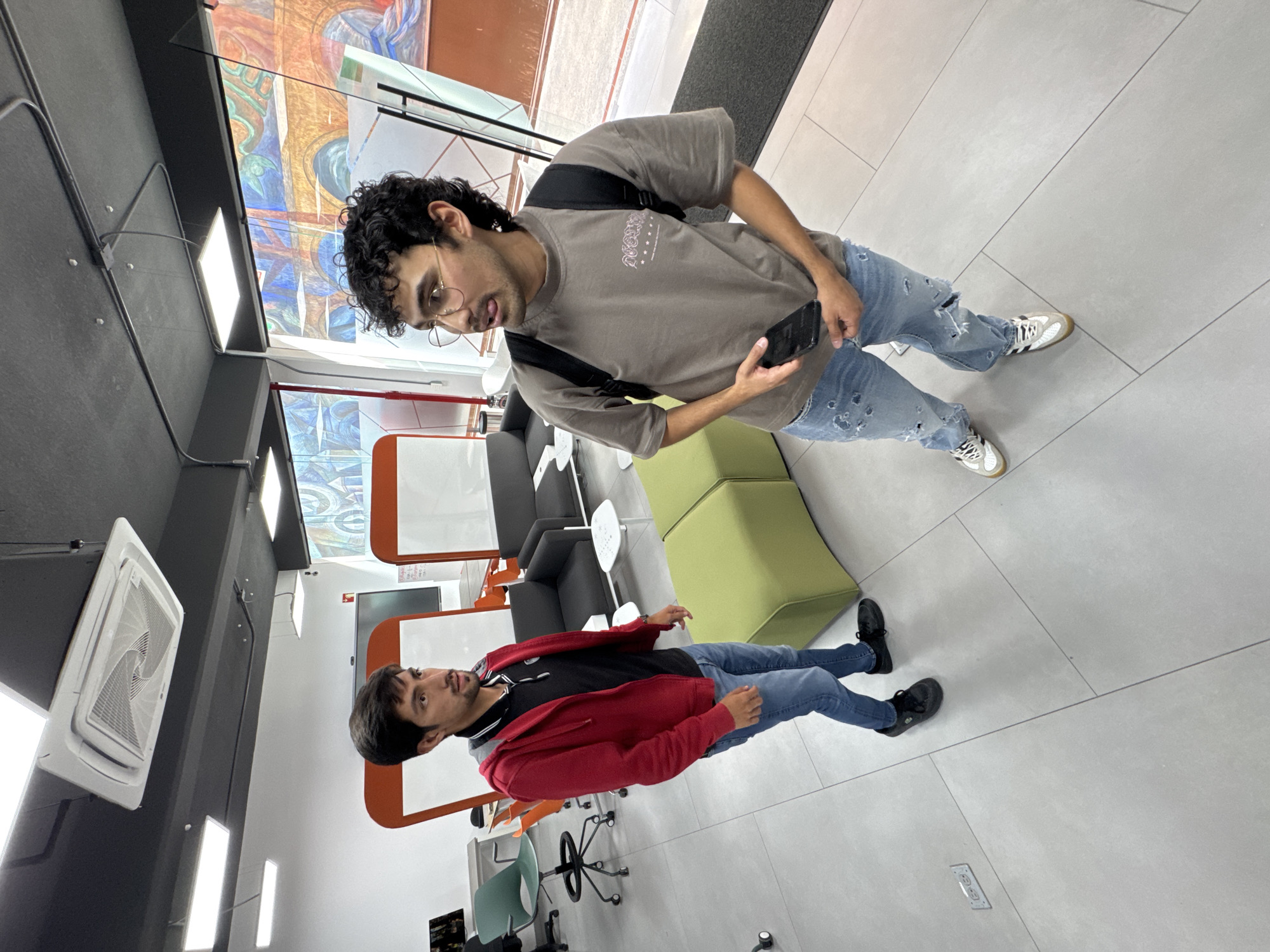 Two men are standing in a modern indoor space. The man on the right has curly dark hair, is wearing glasses, a gray t-shirt, and ripped blue jeans, and is holding a smartphone. The man on the left has dark hair, and a beard, and is wearing a red jacket, a black t-shirt with a white collar, and blue jeans. The location seems to be a public space or office in San Andrés Cholula, Mexico. There are green and black seats nearby.