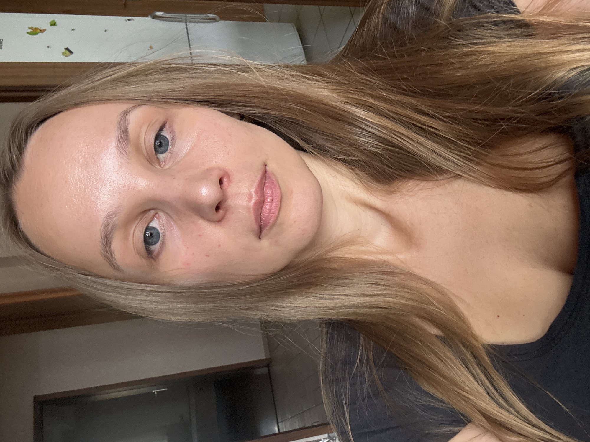 A woman with blonde hair is taking a selfie indoors. She has blue eyes and is looking directly at the camera. Her skin appears fair with some redness and blemishes. She is wearing a dark-colored shirt. The background is blurry, showing a doorway on the left and a white appliance, possibly a refrigerator, on the right. The lighting suggests an indoor environment, likely a home. There are no other individuals or notable objects present. The image does not contain any information that would place it in Überherrn, Germany.