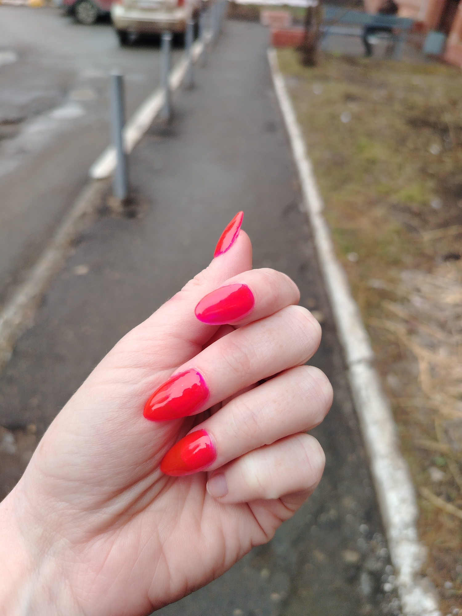 A close-up shot captures a woman's hand with bright red, glossy nail polish. The nails are filed into a long, pointed shape. The background is a blurred cityscape. The ground appears to be wet asphalt, with metal poles lining the sidewalk. Further in the background, there is sparse vegetation and a glimpse of a building. Based on the style of buildings and urban environment, the location is likely in Izhevsk, Russia.