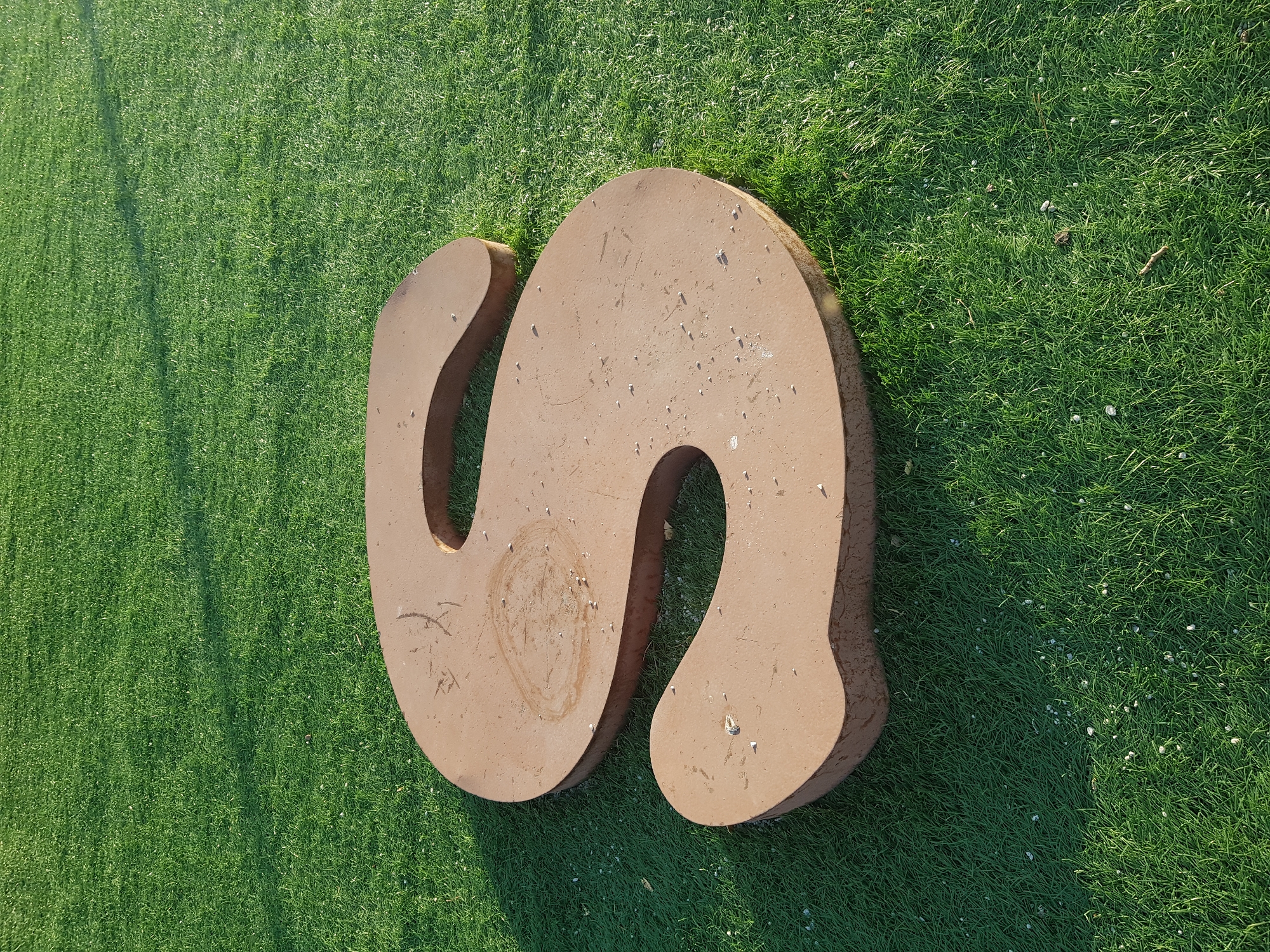 A large, brown, three-dimensional letter "S" made of metal or concrete lies on a bright green, well-maintained lawn. The letter is positioned diagonally across the frame, with its curves facing the viewer. The surface of the "S" appears worn and slightly scuffed, with a circular stain and scattered small white pebbles. The grass is uniformly cut and shows some subtle shadows cast by an unseen light source, possibly the sun. The overall scene is one of an outdoor public space, likely a park or plaza, in the city of Kurchatov, Kazakhstan.