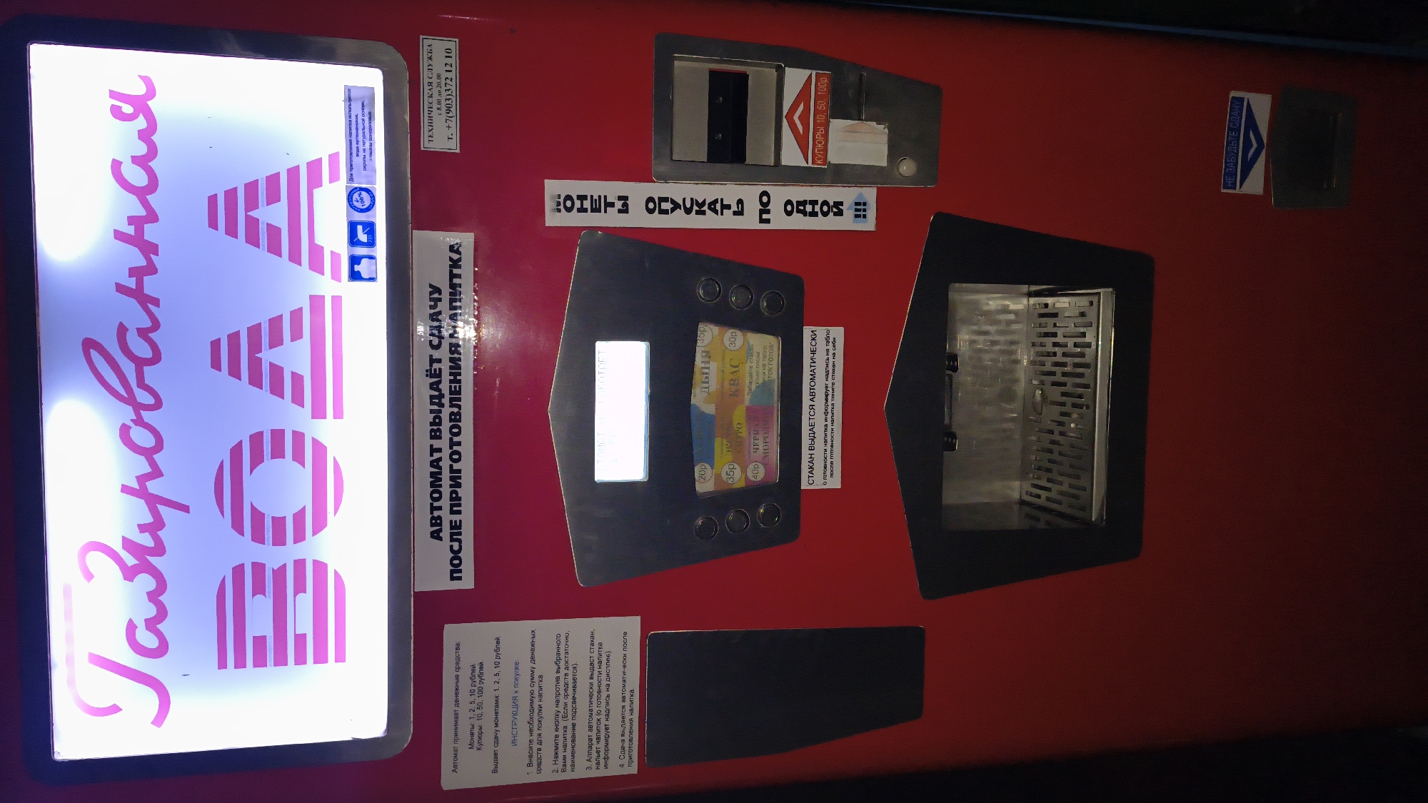 The image shows a red automatic drink vending machine. The machine has a large illuminated sign at the top that reads "Газированная ВОДА" (Carbonated WATER) in pink letters. Below the sign, there is a panel with a small display screen and several buttons and coin slots. Different drink options are listed with prices, such as "ДЫНЯ" (Melon) for 35p, "КВАС" (Kvass) for 30p, "ЧЕРНИКА" (Blueberry) for 40p, and "МОРС" (Morse) for 40p. There is also a cup dispenser and a change return slot. To the right of the main panel, there is a vertical text strip that says "МОНЕТЫ ОПУСКАТЬ ПО ОДНОЙ" (Insert coins one by one) and below it, a slot for coins with prices listed as 10, 50, 100p. At the bottom of the machine, there is another slot labeled "НЕ ЗАБУДЬТЕ СДАЧУ" (Don't forget your change). The machine is situated in an indoor environment. The text in the image indicates that the machine is located in Mikhaylovka, Russia, based on the text on the sign.