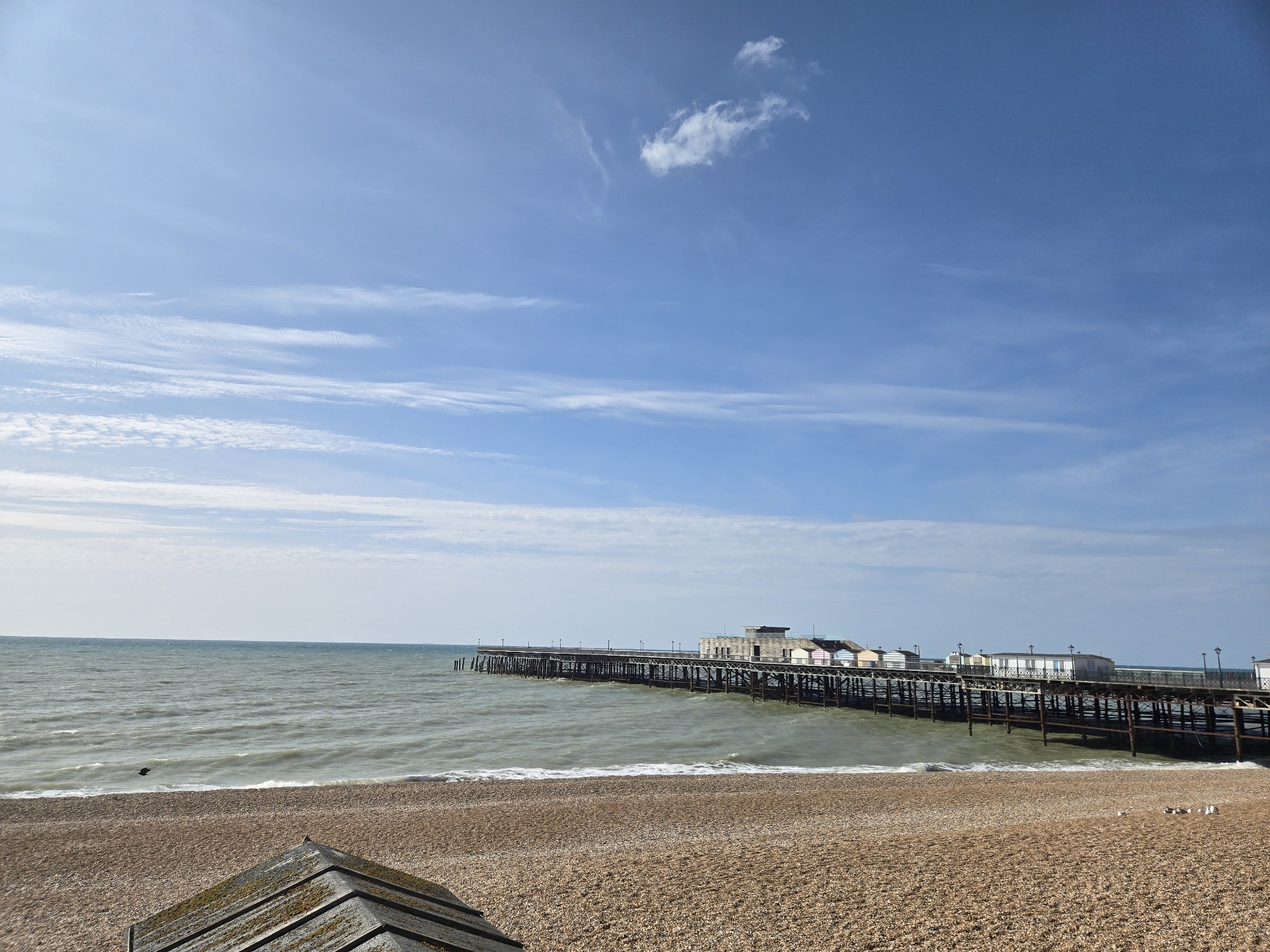 The image shows a view of the St Leonards Pier in St Leonards-on-Sea, East Sussex, United Kingdom. The pier extends out into the sea, with several buildings visible on it, including what appear to be beach huts. The sea is choppy and there are waves breaking on the pebble beach in the foreground. The sky is blue with some white clouds. There are no people visible in the image. A small section of a moss-covered wooden structure is visible in the lower left corner of the image.