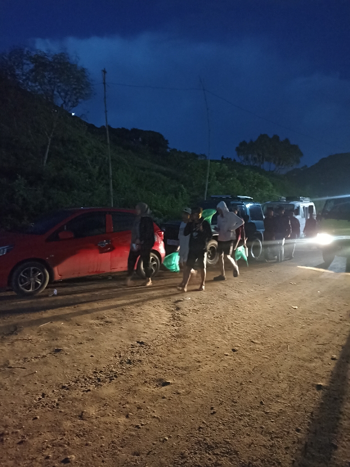 The image captures a group of people and vehicles on a dirt road at night in Kecamatan Pujut, Indonesia. The sky is a deep indigo blue with heavy, dark clouds suggesting either late dusk or early night.
In the foreground, a rough, unpaved dirt road stretches across the frame, illuminated by artificial light sources, primarily headlights from a vehicle on the far right. On the left, a red hatchback car, possibly a Honda Brio, is parked or slowly moving. A group of approximately eight to ten individuals, mostly young adults, are visible walking or standing on the road between the red car and several larger, darker vehicles (SUVs or vans) further down. Many of these individuals are carrying distinctive bright green bags. They are dressed casually, with some wearing hoodies and shorts. Their faces are largely obscured by the low light and motion blur.
Behind the road, a steep hillside densely covered with trees and shrubs rises prominently. Two tall, slender poles, possibly bamboo, are erected on the hillside, with thin lines or wires strung between them, adding to the rural aesthetic. The overall atmosphere is dark, with pockets of light from the vehicles. The activity appears to be a gathering or movement of people, possibly travelers or participants in an outing, along a rural route at night. No discernible text is visible in the image.