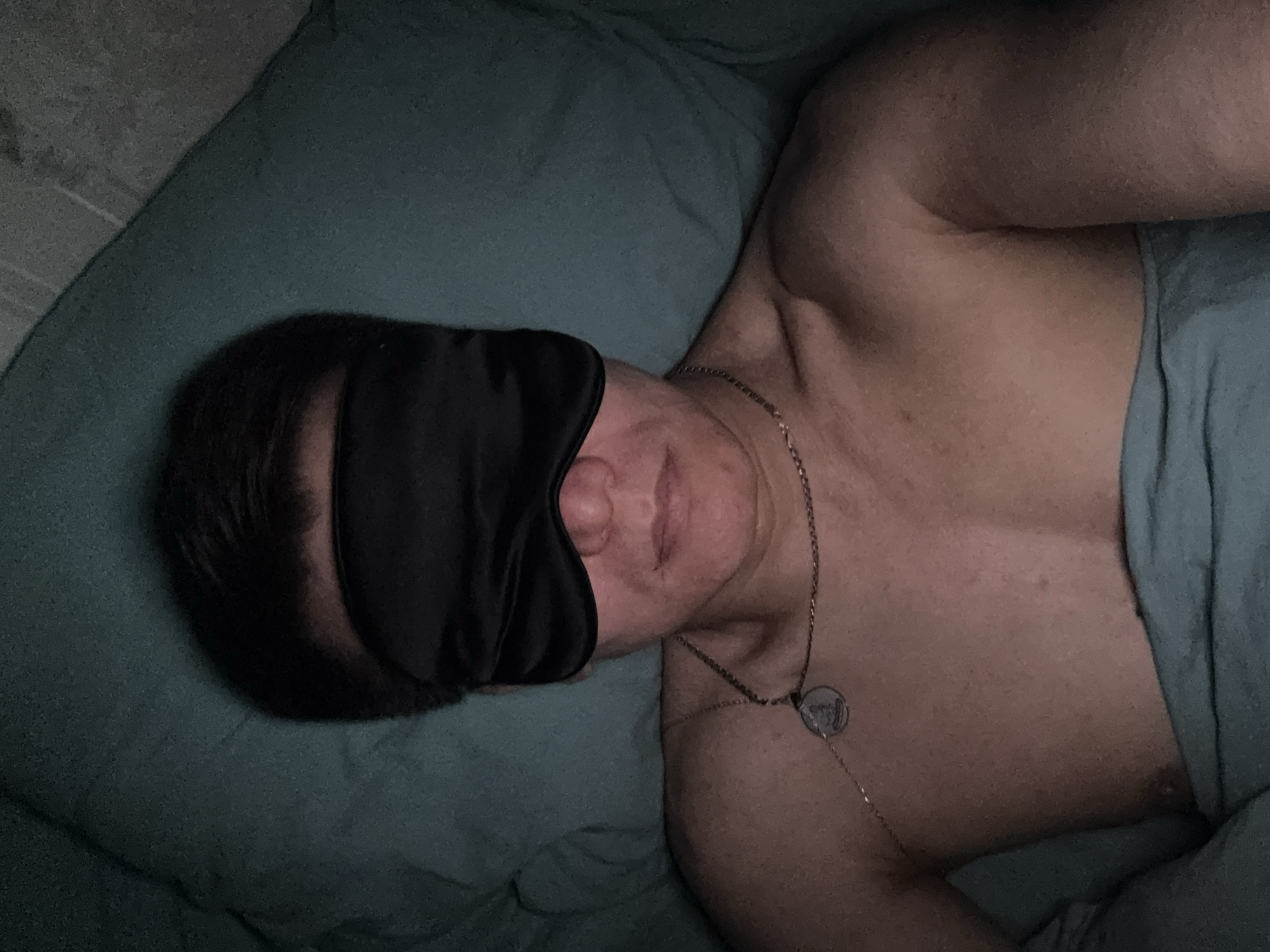 A person wearing a black sleep mask is lying on a pillow under a blanket. The person has dark hair and is wearing a silver necklace with a circular pendant. The scene appears to be indoors and dimly lit.
