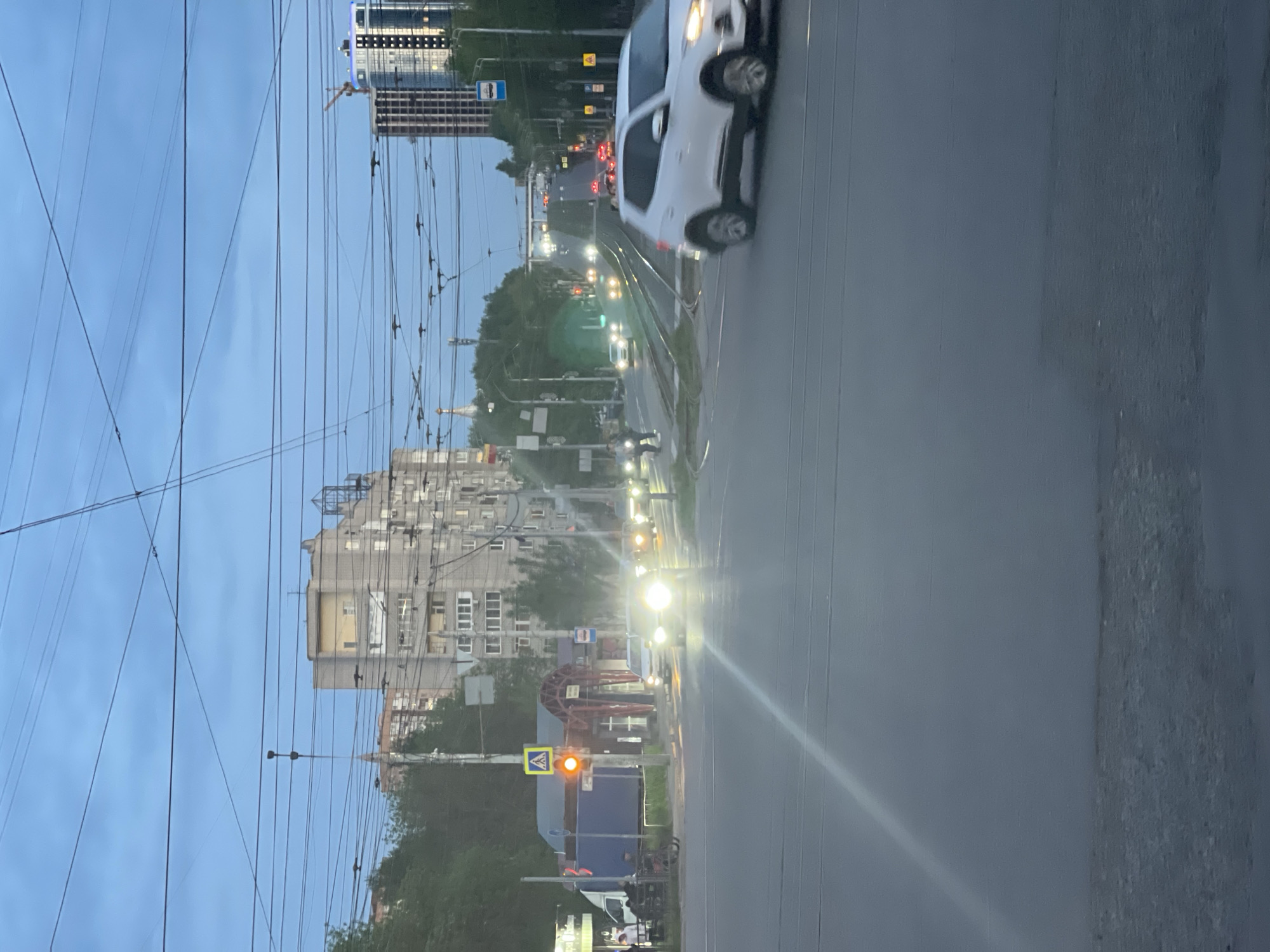 The image depicts a street scene in Izhevsk, Russia. In the foreground, the focus is on the right side of the road, showing a section of the road surface. A blurred white vehicle appears to be in motion in the middle of the image, on the road with tram tracks. Further down the road, traffic and streetlights are visible. On the left side, there is a large apartment building, overhead electrical wires, and various street signs. A pedestrian crossing sign is illuminated with an orange light. The sky is overcast.