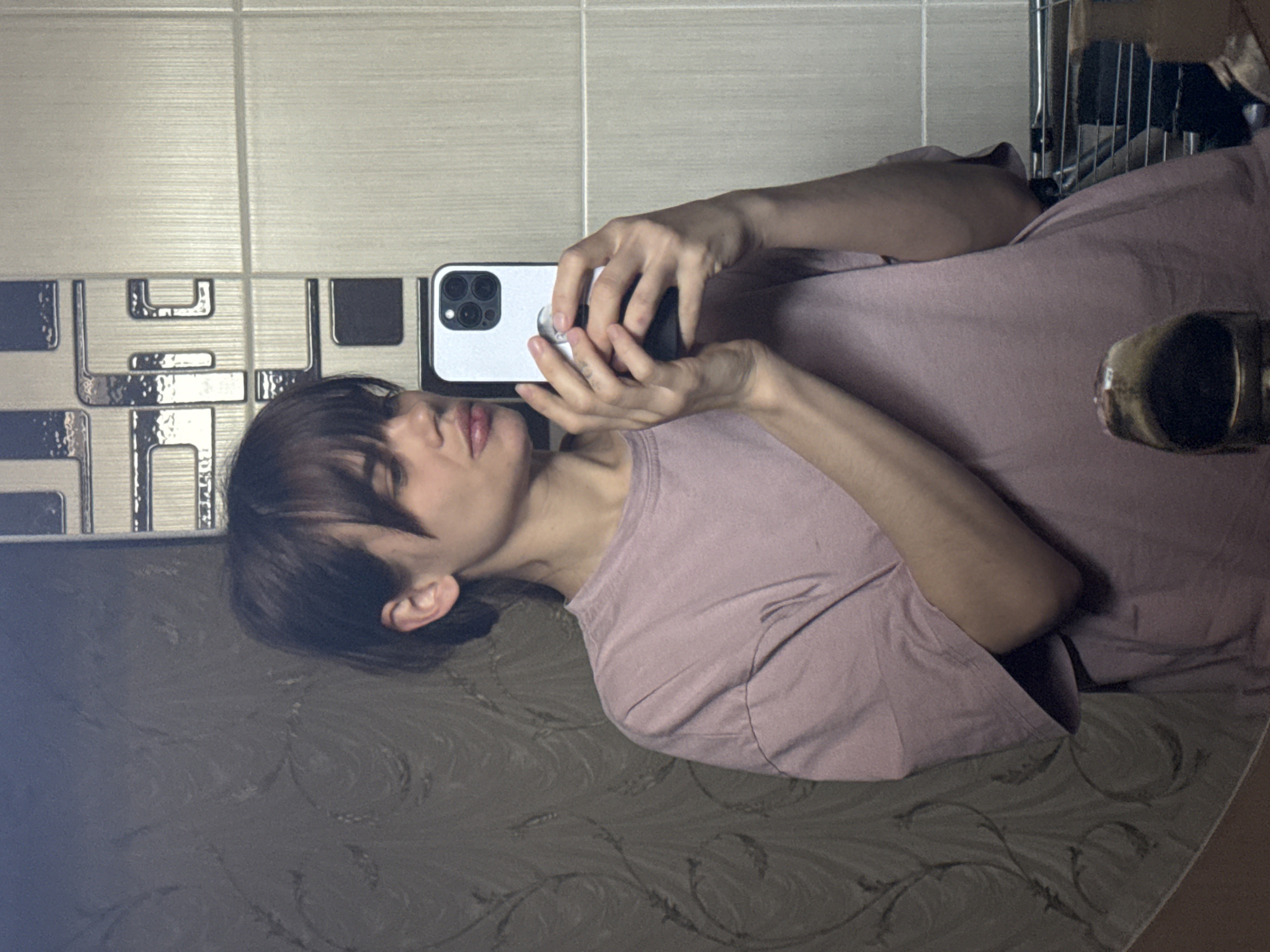 A person with dark hair is taking a selfie with a white phone in what appears to be a bathroom. The walls are tiled with a combination of light and dark colored tiles with a geometric pattern. The person is wearing a pink t-shirt. Part of a shelf with items is visible in the upper right corner of the image. The person is lying on a surface with a dark, patterned cover. There is no definitive information in the image to confirm its location as Rostov-on-Don, Russia.