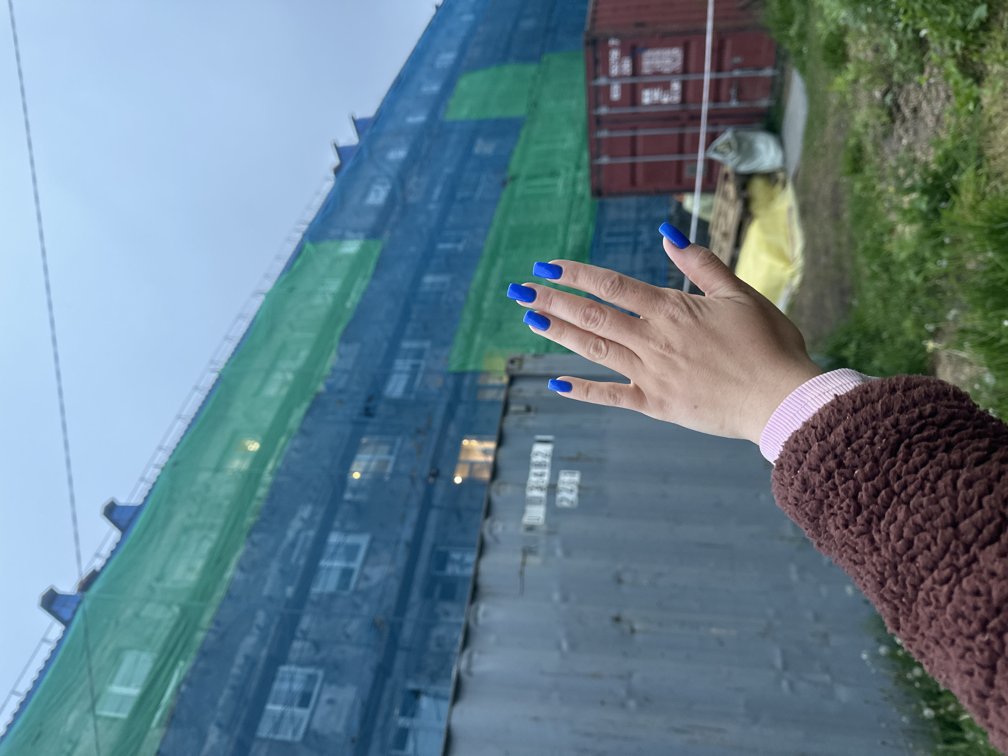 An outdoor medium shot captures a person's hand with bright blue fingernails extended towards a large building covered in blue and green netting. The netting obscures most of the building's facade, but glimpses of windows and lights are visible through the mesh. In the foreground, a corrugated metal shipping container with faded markings stands to the left of the hand. To the right, another red shipping container is partially visible, along with some construction materials, including a yellow tarp and a pallet. The background shows a slightly overcast sky and an area of overgrown grass. The scene appears to be an urban or industrial area, possibly undergoing construction or renovation, in Vilyuchinsk, Russia. The person is wearing a brown fuzzy jacket with a pink cuff visible at the wrist. The hand is positioned in a relaxed, open posture, with fingers slightly splayed.