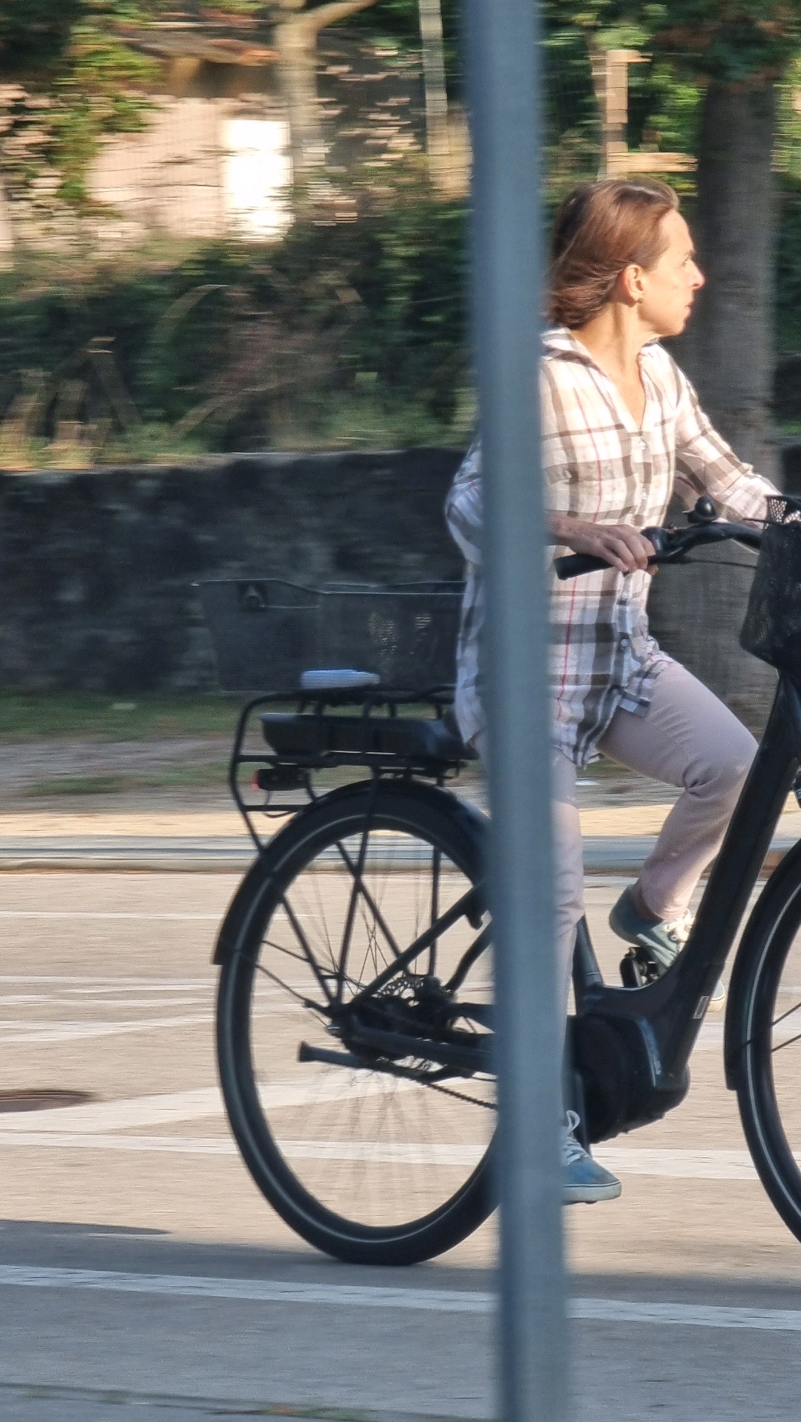 A person is riding an electric bicycle on a paved surface, possibly a road or pathway, with a blurred background of trees and structures. The person is wearing a plaid shirt and light-colored pants. The bicycle has a basket on the rear and appears to be in motion. The lighting suggests it is daytime. The location is identified as Sant Joan les Fonts, Spain.