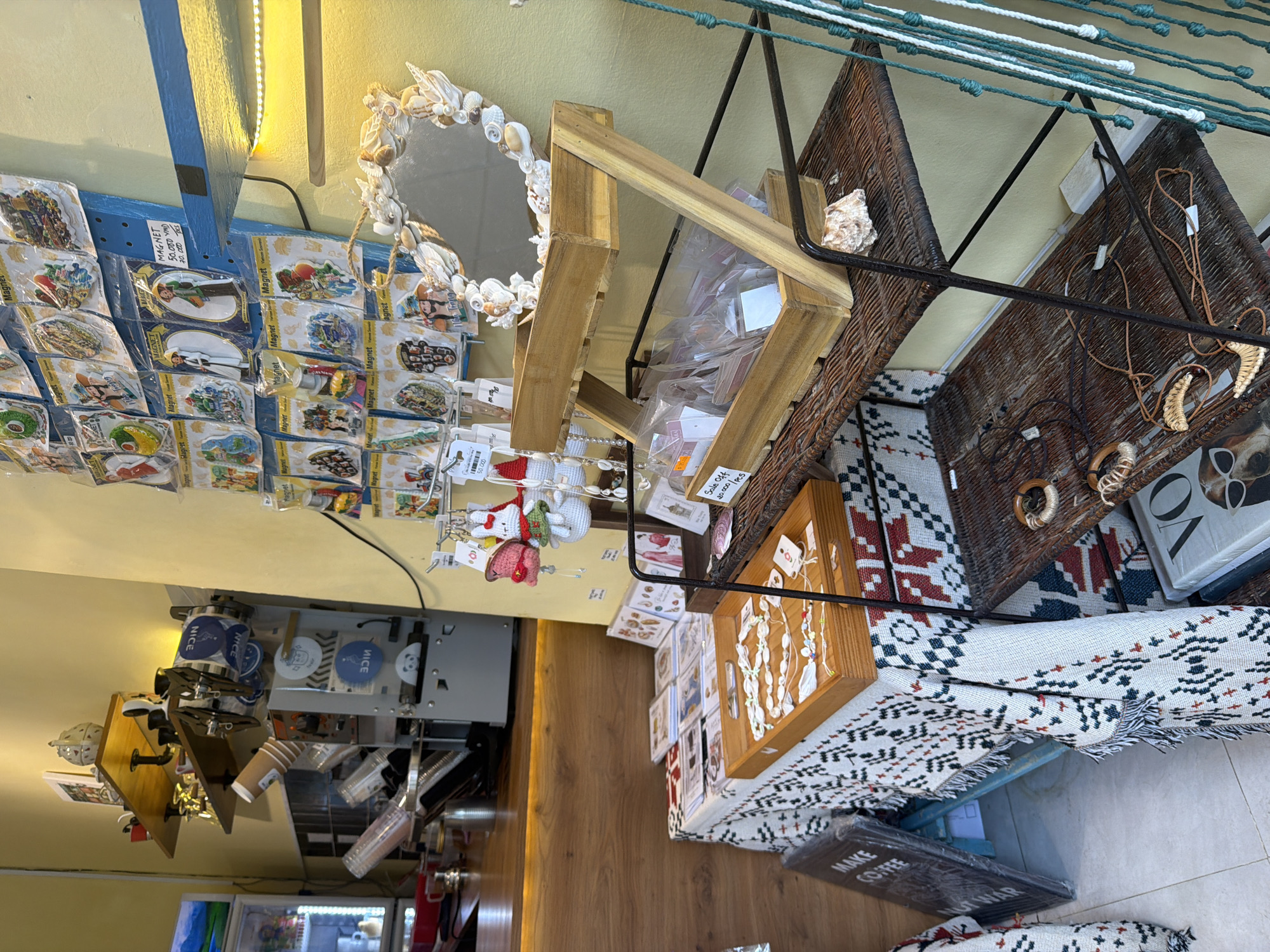 The image captures a cluttered interior space in Bac Nha Trang Ward, Vietnam. Merchandise is densely packed: magnets hang from a display, a mirror framed with seashells sits nearby, and wooden shelves hold various items. Woven baskets containing jewelry and decorative elements rest atop a patterned blanket. A wooden display case showcases more jewelry, and a sign reading "MAKE COFFEE NOT WAR" leans against the wall. In the background, a coffee machine and cups suggest the space may be a cafe or a shop combined with a cafe.
