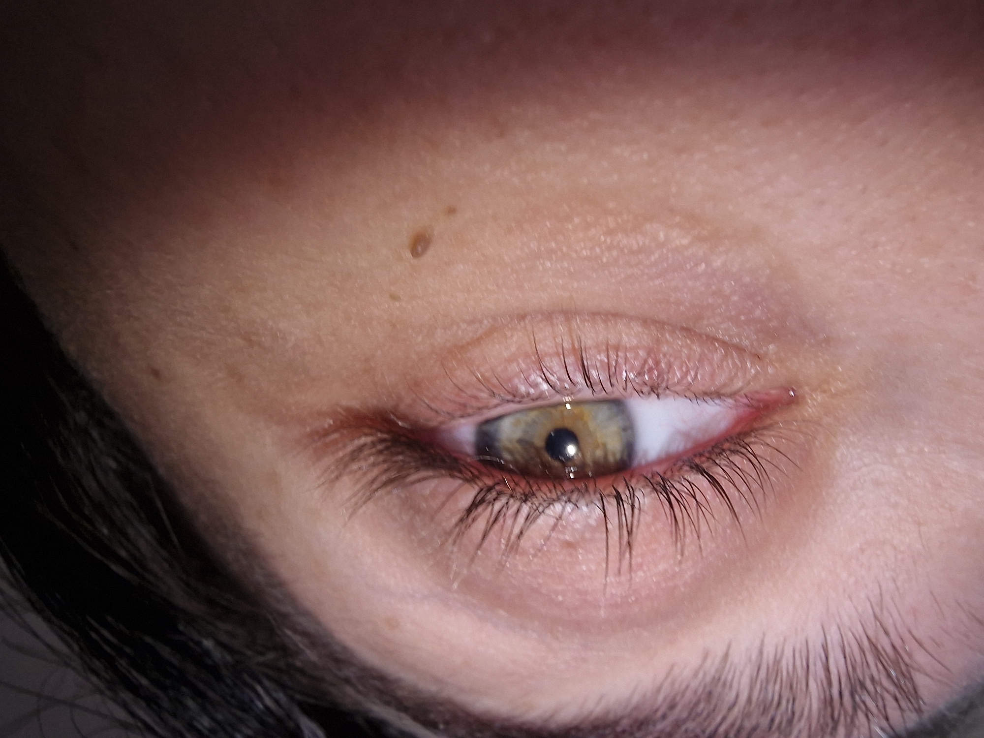 The image is a close-up of a human eye, specifically the right eye, with a light complexion, dark eyelashes, and a hazel-colored iris. There is a small mole visible above the eyelid. Hair is visible near the bottom edge. The surrounding skin shows texture and some lines. The location is unspecified.