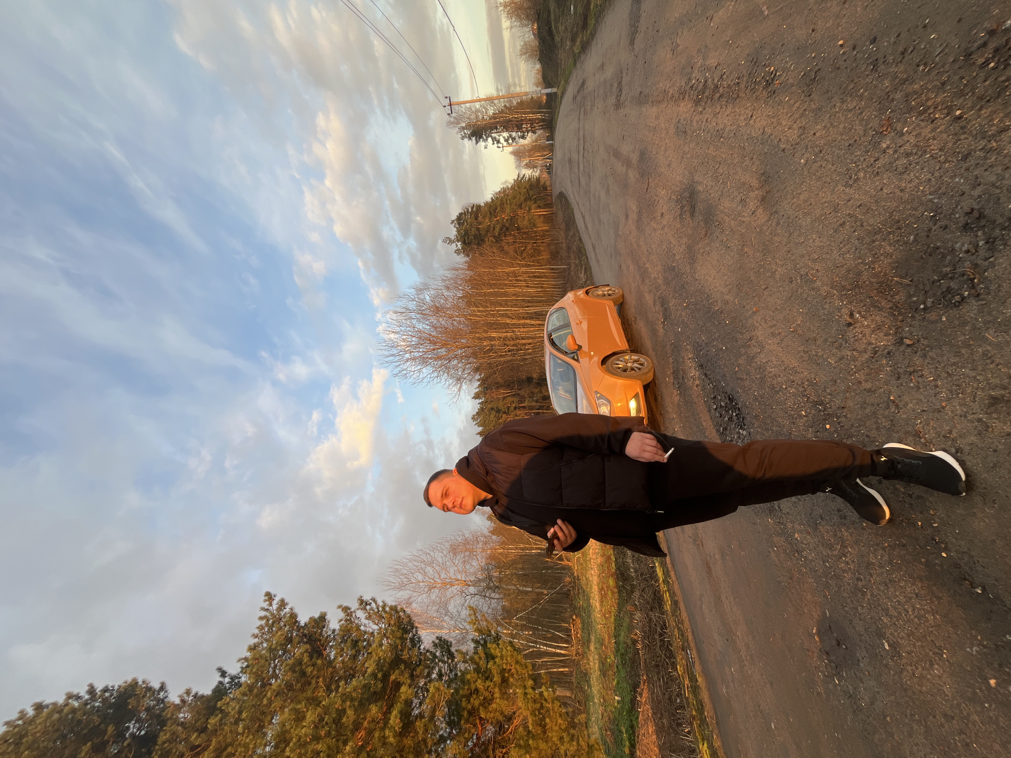 The image captures an outdoor scene at golden hour, characterized by warm, low-angle sunlight casting long shadows and illuminating clouds with soft orange and blue hues. The primary subject is a young man, standing on a dirt road and looking towards the viewer. He wears a black padded jacket, black trousers, and black sneakers with white soles. He holds a lit cigarette in his right hand and a mobile phone in his left.
Beside the man and slightly behind him is a bright orange hatchback car, parked on the unpaved road. The road surface is composed of dirt and small stones, suggesting a rural or semi-rural environment. The background features a landscape with various trees, including bare deciduous trees and green conifers, indicating either late autumn or early spring. Overhead, power lines are visible against the cloudy sky. The overall setting is consistent with an outdoor location in Voronino, Russia, with no discernible text visible in the frame.
