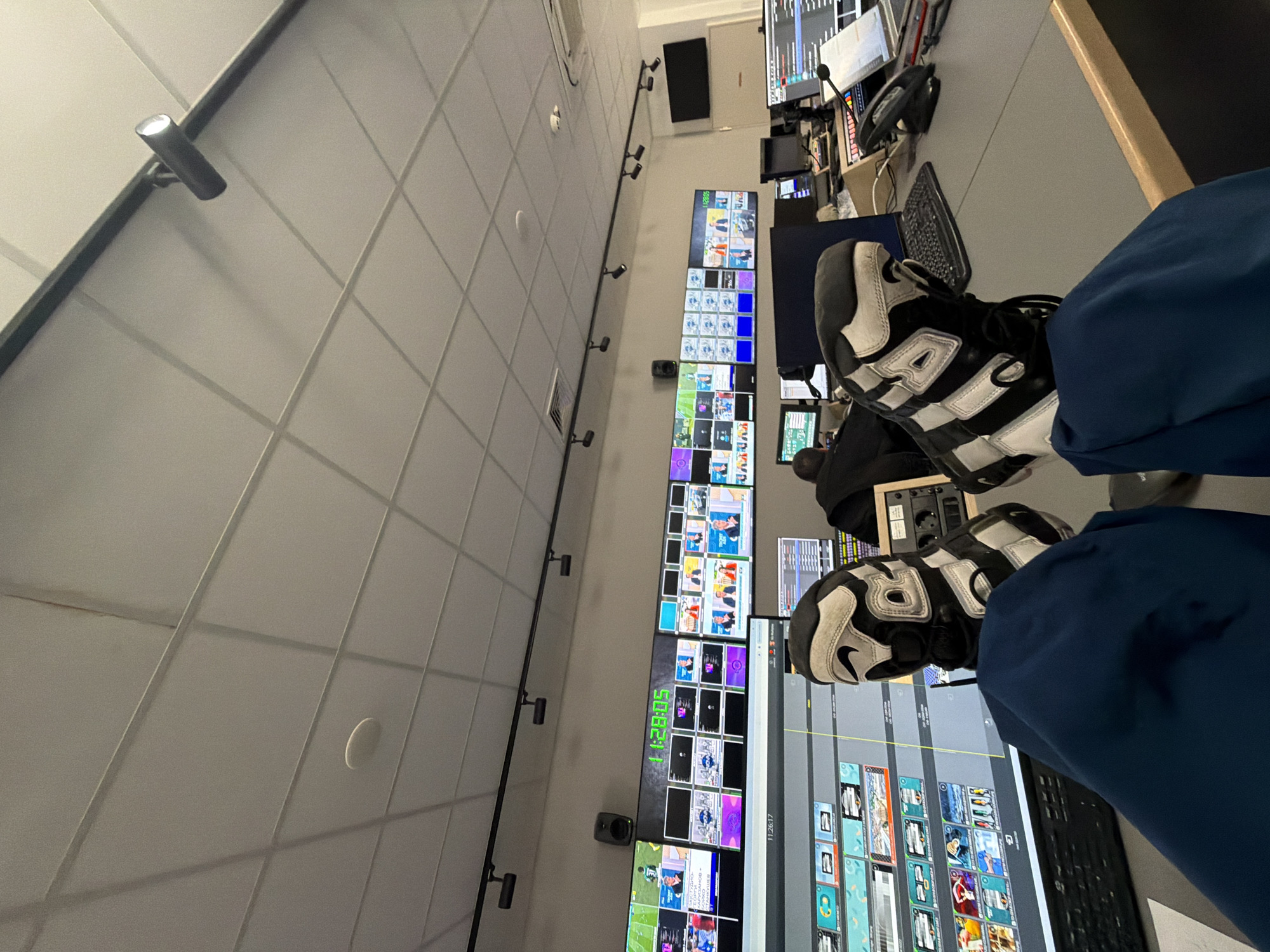The image shows a person sitting with their feet on a desk in a room with multiple monitors displaying various images and data. The person is wearing blue pants and black and white sneakers. The room appears to be a control or monitoring room, possibly for a broadcast or security operation, given the number of screens. There is also a row of spotlight-style lights attached to a rail on the wall. There is no information to identify the location as Sofia, Bulgaria, or any location at all.