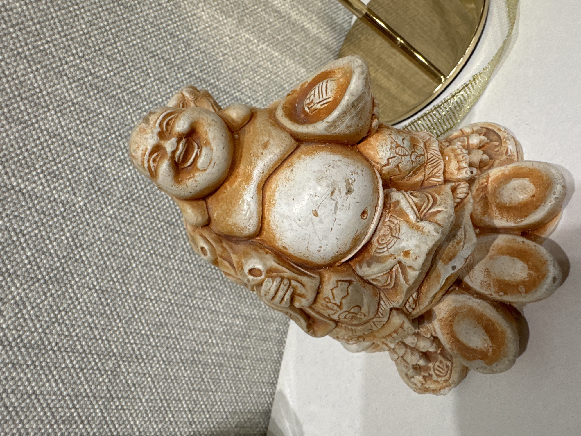The image features a detailed, high-angle view of a decorative figurine, likely representing a Laughing Buddha or Hotei. The figurine is made of a light beige or off-white material, accented with reddish-brown patination that highlights its textured surface and carved details. It portrays a figure with a plump body, a prominent round belly, and a serene, smiling face with closed eyes.
The figurine appears to be holding several objects, possibly ingots or coins, symbolizing prosperity. Some of these objects feature faint, stylized vertical markings that resemble characters or symbols, though they are not clearly decipherable. Intricate patterns and textures adorn the figure's clothing and the items it carries, suggesting traditional Asian craftsmanship.
The figurine rests primarily on a clean white surface, possibly a table or shelf. To the left, a textured grey fabric, characterized by a fine, repetitive weave pattern, is visible. In the background, partially visible on the right, is a metallic gold object, possibly part of a lamp or another decorative item, accompanied by a thin, glittery gold ribbon or trim.
The scene is a still life, showcasing the figurine as a decorative art piece, with no explicit activity or event taking place. The lighting is bright and even, typical of an indoor setting, without indicating a specific time of day. The figurine's surface shows variations in color and texture, suggesting either age or an intentional antique finish. There are no visual cues within the image itself to confirm the location as Chaikovsky, Russia.