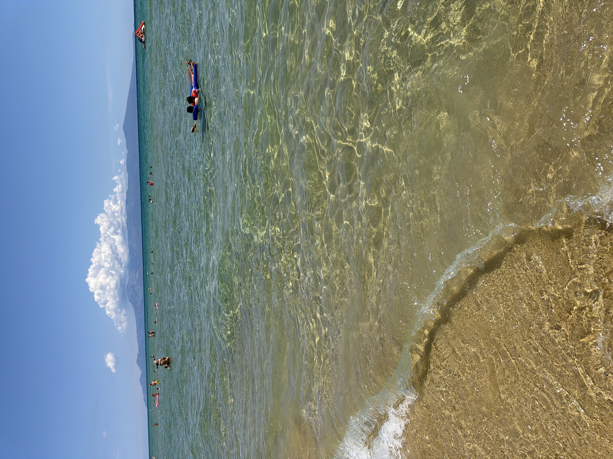 The image captures a sunny day at the beach in Nea Peramos, Greece, with clear, shallow water and a sandy shore. In the foreground, a gentle wave breaks on the golden sand, its translucent water revealing the textured seabed. The middle ground is populated by numerous people enjoying the sea. Some are swimming or wading, while others are relaxing on inflatable devices. Two individuals are visible on a blue inflatable raft in the middle of the frame. Further in the distance, the sea extends to a hazy coastline with mountains under a bright blue sky dotted with white clouds. The overall scene depicts a leisurely and idyllic beach day.