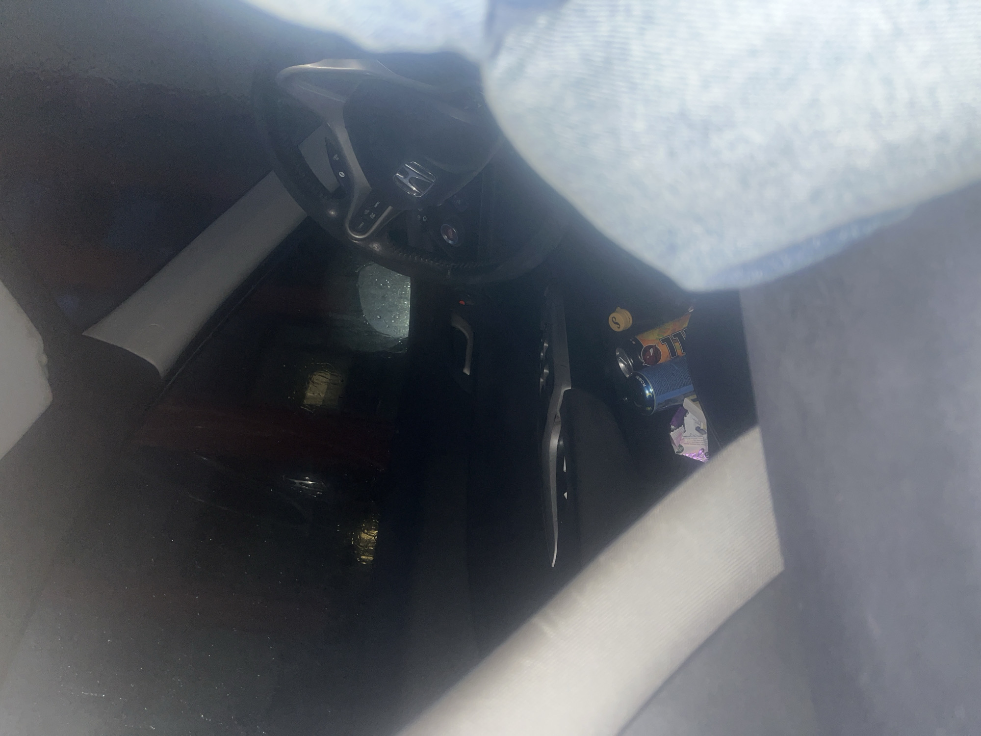 The image offers a high-angle, downward view into the interior of a car, characterized by dim lighting, suggesting it might be evening, night, or an overcast day. The primary subjects are the car's internal components and miscellaneous items.
The steering wheel, black with visible control buttons on its spokes, is prominently featured. A clear "Honda" logo is centered on the steering wheel. Below it, a section of the dashboard and what appears to be a push-button start mechanism are visible. The driver's side floor is dark and shows some reflective qualities, potentially indicating a wet surface.
On the passenger side floor or in an adjacent storage area, several objects are present. These include multiple cans, with one blue/purple can partially showing the brand "MONSTER" and other text, possibly "LITT" on another can. A yellow bottle cap with the letter "S" is also visible, alongside a small, crumpled white and purple plastic wrapper, likely from a snack.
A significant portion of the top and right side of the image is covered by a light blue, textured fabric, strongly resembling denim, which is likely part of the clothing of a person (e.g., a leg or jacket). A lighter grey or white fabric is visible in the bottom right, possibly a seatbelt or part of the car seat. No people are directly visible, and no specific activity is taking place within the stationary vehicle. The image does not contain any direct location cues pertaining to Varna, Bulgaria.
