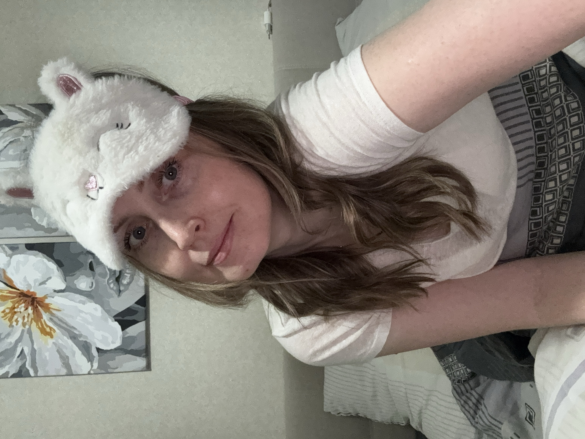 This medium close-up selfie, taken indoors in Chaikovsky, Russia, features a young woman lying down and looking towards the camera. She has long, wavy brown hair, blue-grey eyes, and fair skin with some freckles, displaying a soft, relaxed expression with a slight smile.
She is wearing a simple white t-shirt and a novelty white, fluffy sleep mask, shaped like a cat or rabbit with pink-lined ears and embroidered closed eyes, which rests on top of her head.
The background reveals a bed with patterned and striped bedding in grey and white tones. To her left, a framed piece of art hangs on the wall, showcasing a stylized white flower with a yellow-orange center against a grayscale abstract background. An electrical cord or charger is partially visible on the wall behind her head. The lighting is artificial, contributing to a cozy and relaxed indoor atmosphere, possibly in the evening. No text is visible in the image.