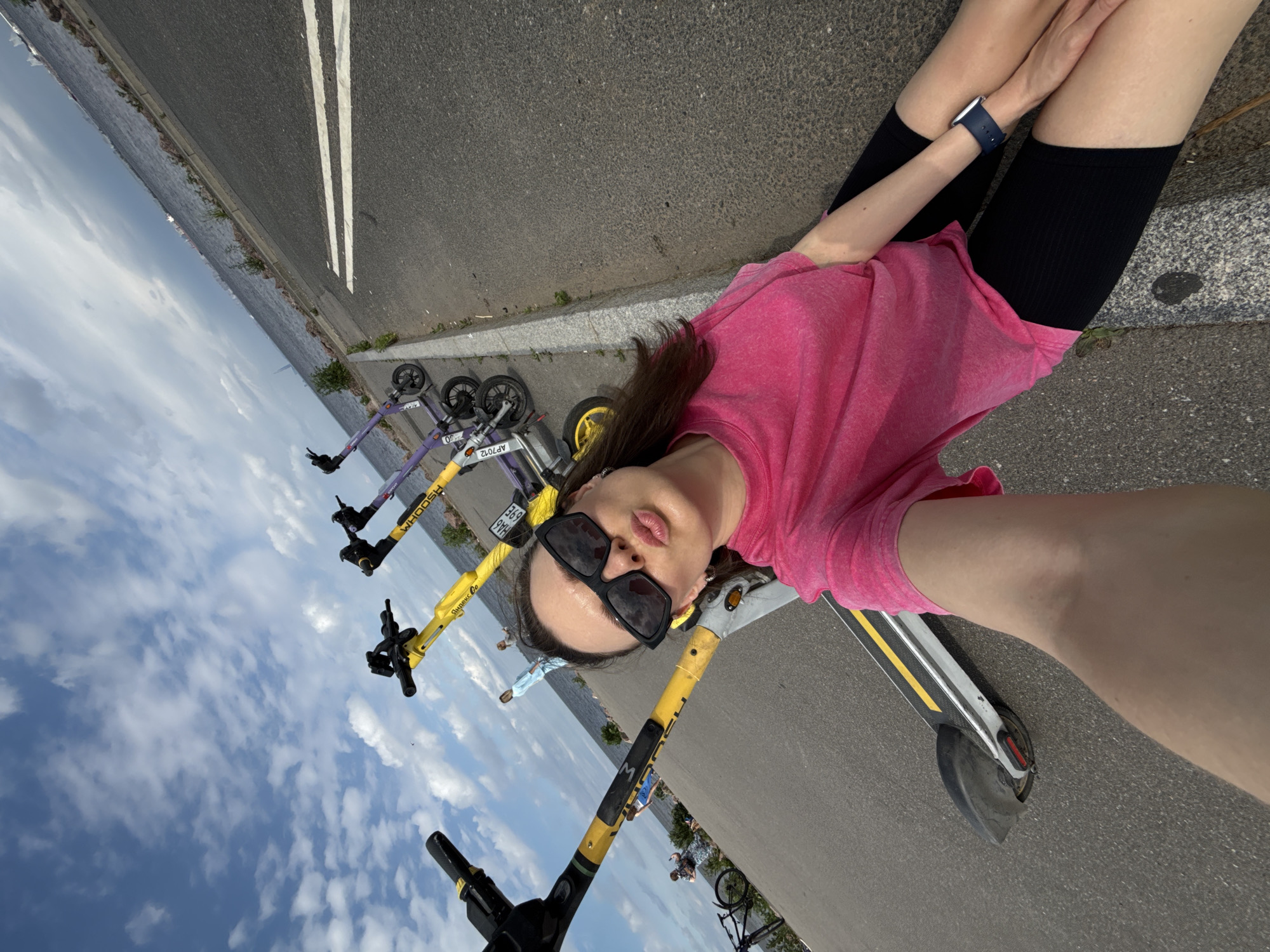A woman is taking a selfie while sitting on the ground next to a row of electric scooters. She is wearing a pink t-shirt and black shorts, with large black sunglasses and a blue watch. The scooters are parked along a paved path next to a body of water. The sky is cloudy, and there are some buildings in the distance across the water. The scene appears to be in Saint Petersburg, Russia.