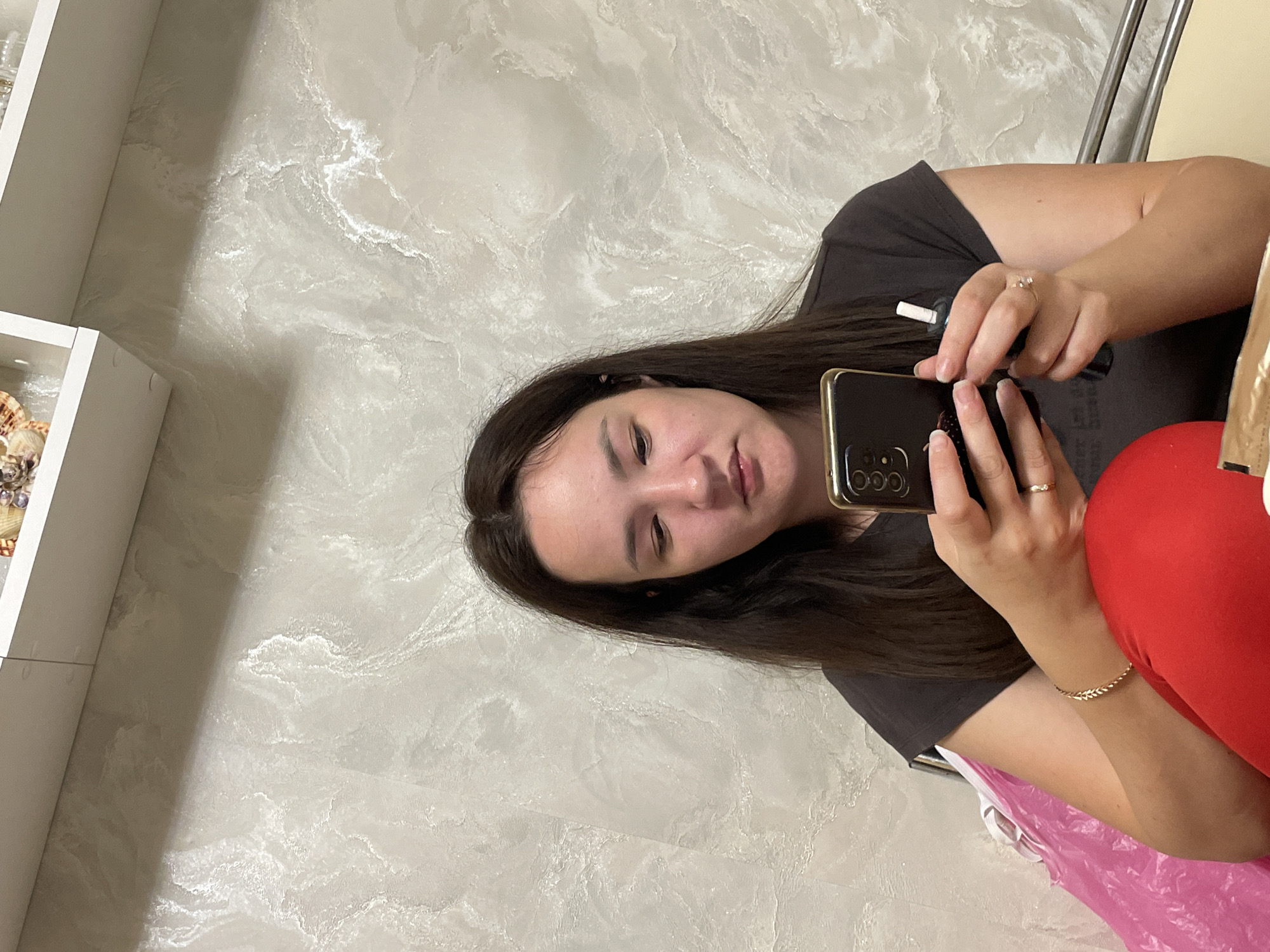 A young woman with long, dark hair is sitting and looking at her phone. She is wearing a dark t-shirt and red pants. She has a gold bracelet on her left wrist and a ring on her left ring finger. She is holding a dark-colored phone in her hands. In her right hand, she is holding a white and black cylindrical object. A pink plastic bag is visible to her left. The background is a textured wall with a shelf above it. The location is not specified in the image.