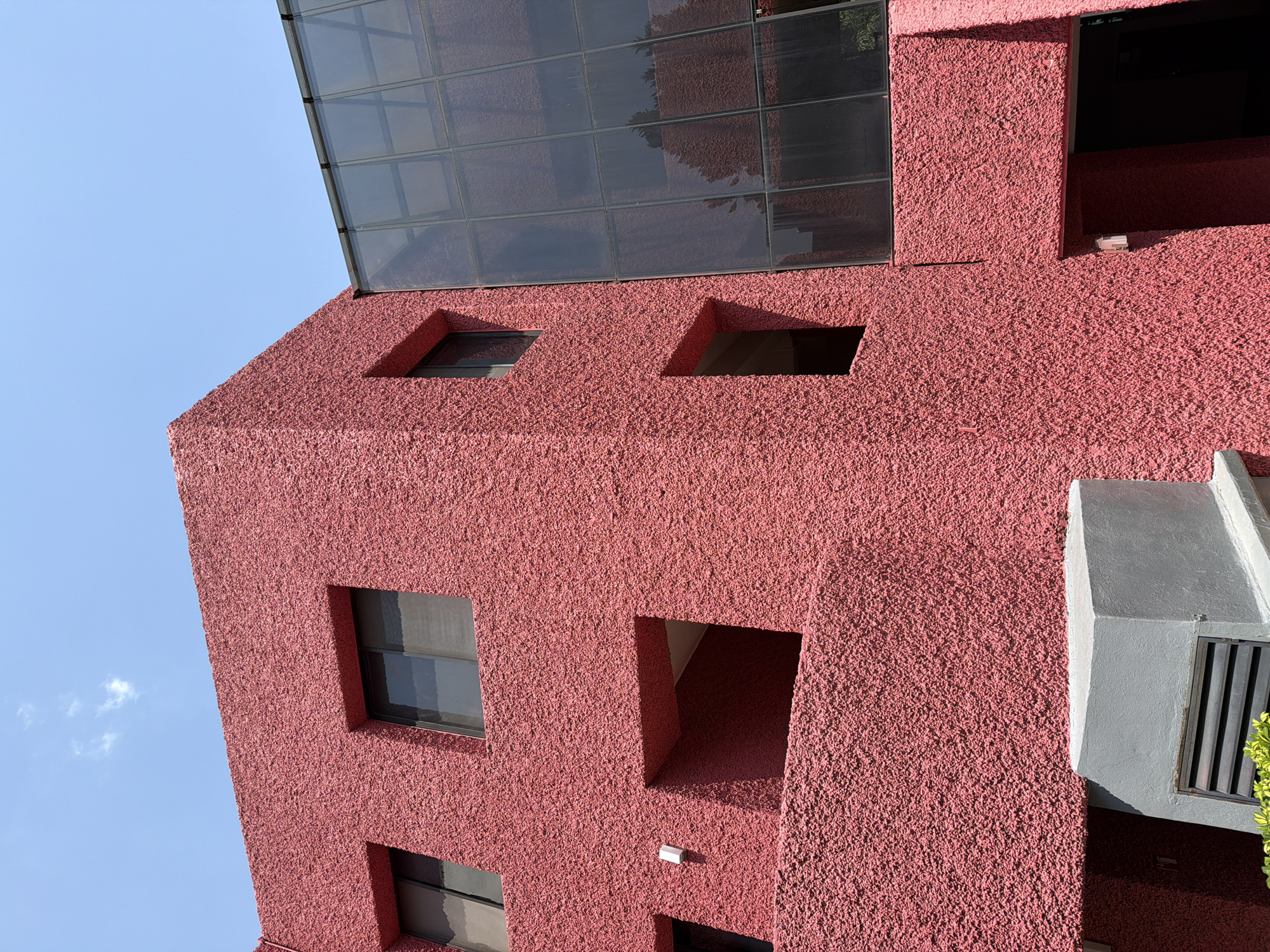 An upward view of a red-plastered building with square window openings, located in San Andrés Cholula, Mexico. The building has a glass-paneled structure at the top. The sky is light blue. A portion of an air conditioning unit or similar ventilation fixture is visible on the right side.