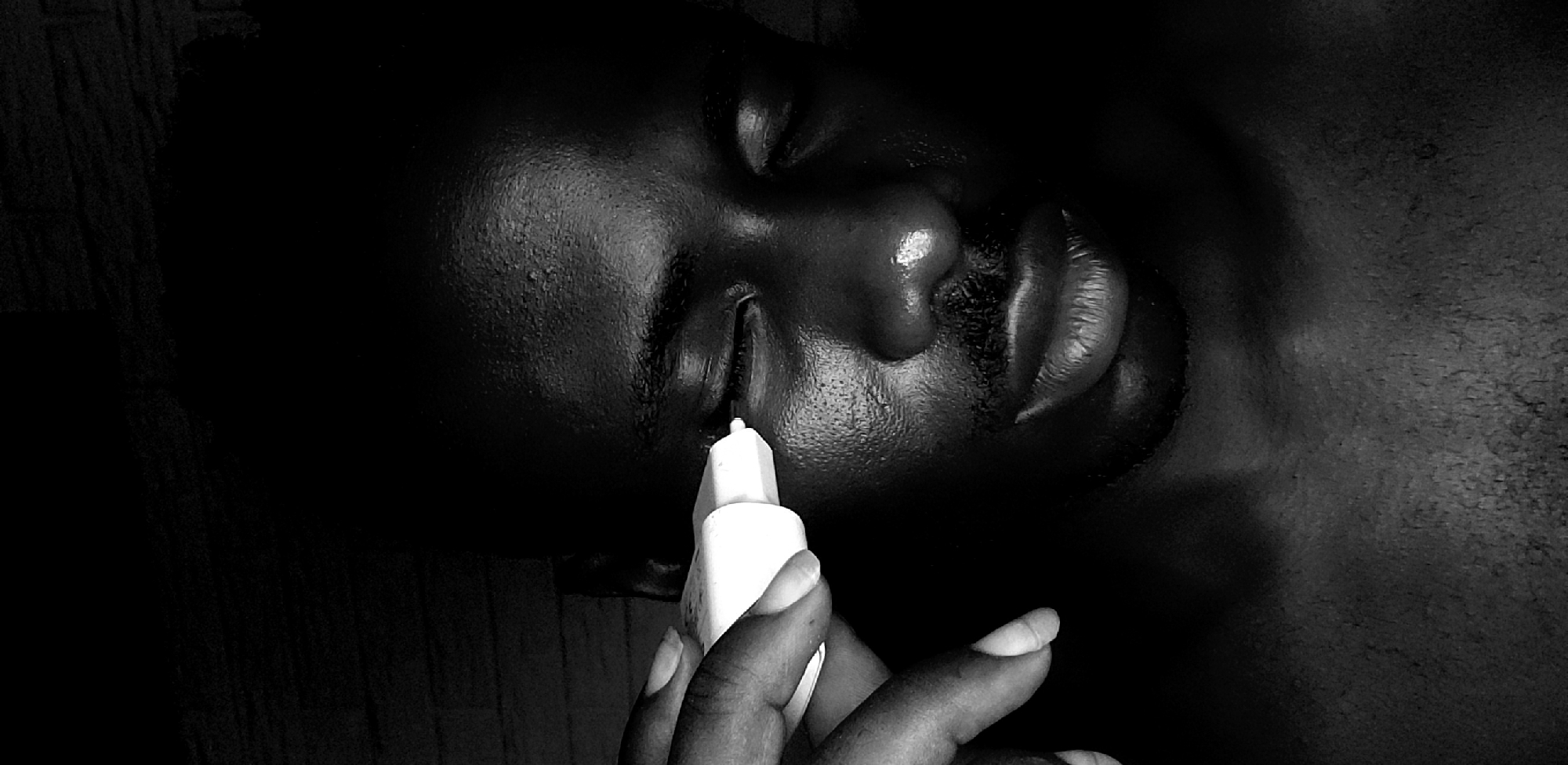 A close-up, black and white photograph shows a person lying on their side, eyes closed. A hand holds a white bottle near the person's eye, possibly administering eye drops. The person has dark skin. The background is dark and indistinct. The scene is not identifiable as being in Narayi, Nigeria, or any specific location.