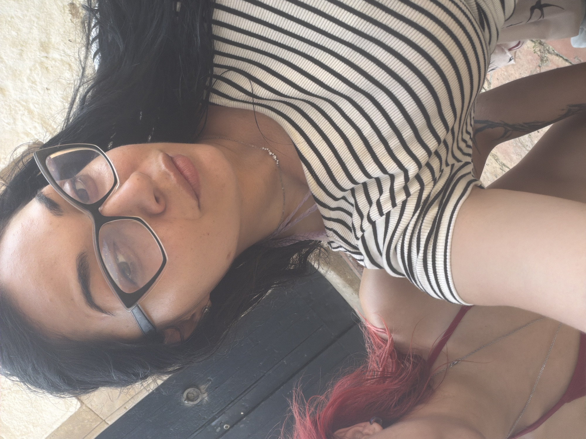 The image shows a close-up of two women. The woman in the foreground has long, dark hair and is wearing cat-eye glasses, a striped shirt, and a delicate necklace. The woman in the background has bright red hair and is wearing a red top and a silver necklace. They appear to be outdoors, with a textured wall in the background and a cobblestone-like surface beneath them. The scene is brightly lit, suggesting it is daytime. There is no specific information in the image to suggest a location in Blanes, Spain.