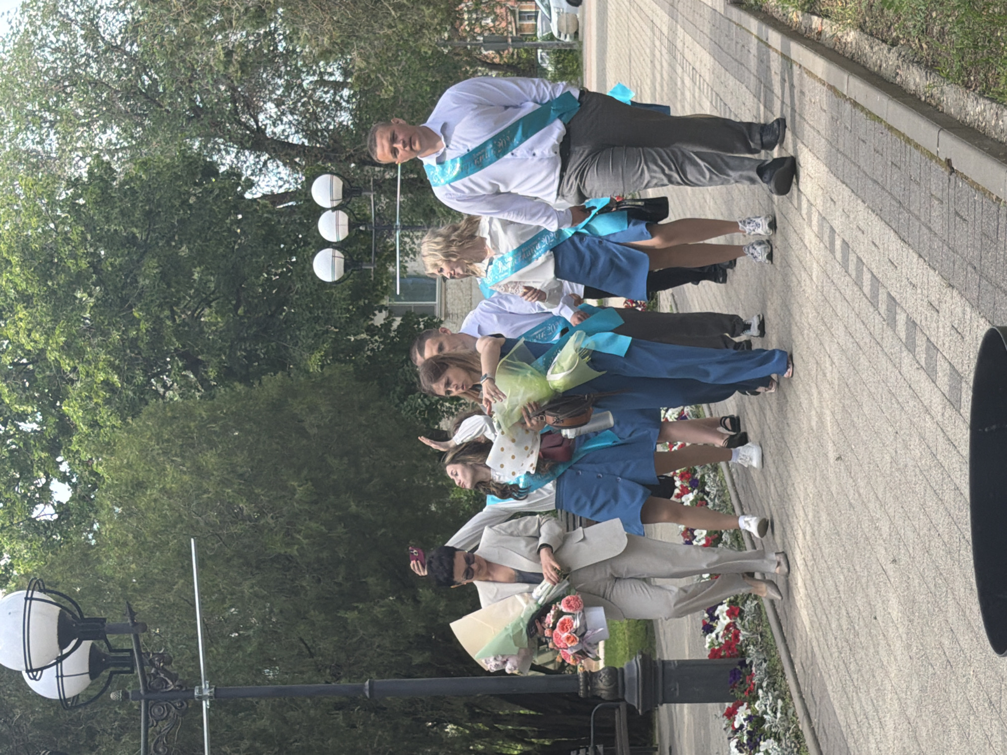 A group of people are walking on a paved sidewalk in an outdoor setting, likely in the city of Leningradskaya, Russia. The group consists of both men and women of varying ages. They are wearing sashes, suggesting they may be graduates. Some individuals are holding bouquets of flowers. One person appears to be taking a photo with a mobile phone. The environment includes trees, lampposts, and flower beds along the sidewalk. Buildings are visible in the background.