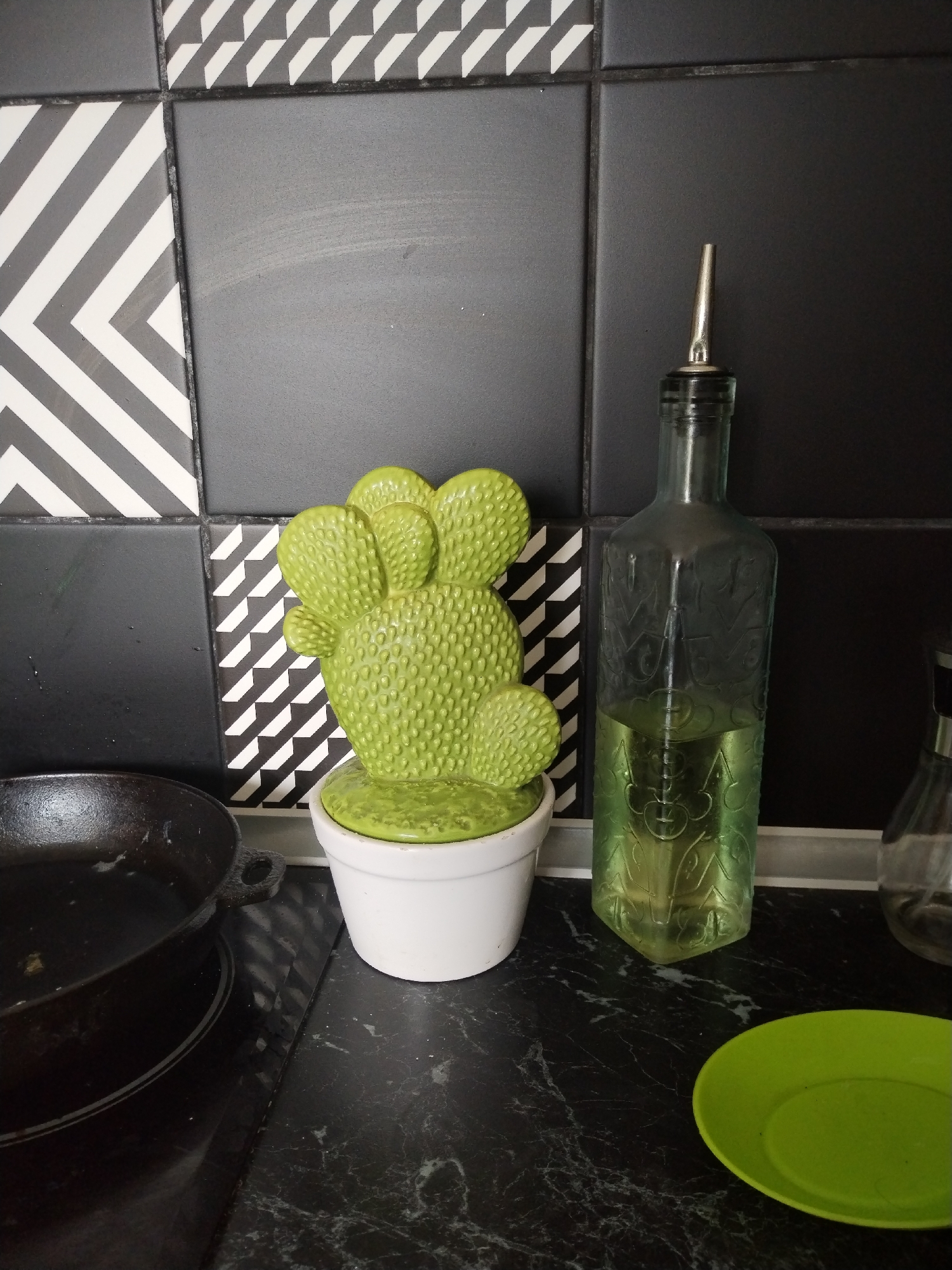 The image presents a close-up view of a contemporary kitchen countertop and backsplash, dominated by a black and white color scheme with vibrant green accents. The background features a striking backsplash composed of large tiles: some are solid black, while others display bold black and white geometric patterns, including chevron or herringbone designs.
On the left side, a black cast iron pan rests on what appears to be a dark, reflective stovetop surface. Adjacent to it, near the center of the frame, is a decorative, bright lime green cactus-shaped object, possibly a pot holder or trivet, housed within a simple white cylindrical pot. The cactus has a distinctive textured, bumpy surface.
To the right of the cactus, a clear glass bottle, fitted with a silver-colored metal pourer spout, stands tall. The bottle contains a light green liquid, likely olive oil, filling about a third of its volume. The glass of the bottle is embossed with subtle decorative patterns. A smaller, clear glass shaker, possibly for salt or pepper, is partially visible behind and to the right of the oil bottle.
The dark countertop beneath the objects features a marble-like pattern with subtle white veins. In the lower right corner, a bright lime green, circular saucer or small plate is placed on the counter. The scene is well-lit, typical of an indoor setting, but the specific time of day is not discernible. No people are visible, and there is no discernible text within the image.
