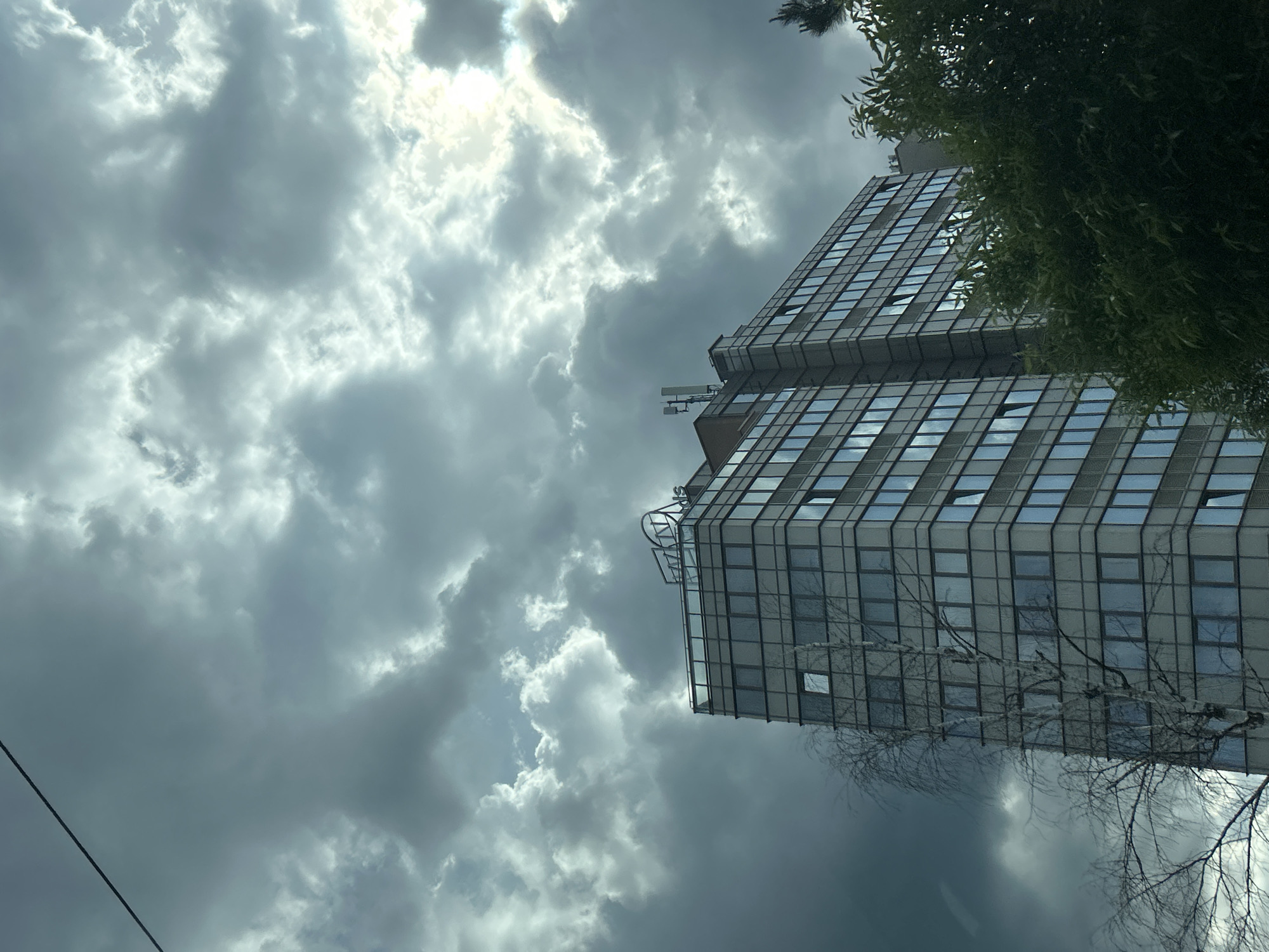 A low-angle shot reveals a tall, modern building with a grid of windows set against a dramatic, cloudy sky. The building, appearing in the city of Sofia, Bulgaria, has a gray facade and a multi-tiered structure. Some windows are open. Foliage partially obscures the right side of the building, while bare tree branches are visible at the bottom. The sky is filled with dark, ominous clouds, with patches of brighter light breaking through. A power line cuts across the lower left corner of the image. The photo appears to have been taken on a day with unsettled weather conditions.