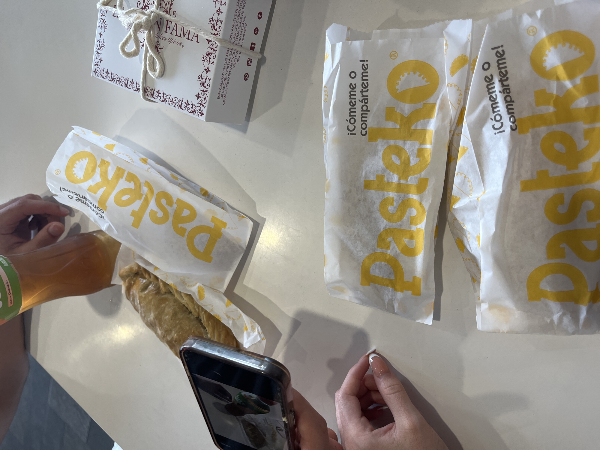 The image depicts a casual indoor setting, likely a tabletop, featuring various food items and human interaction. Two pairs of hands are visible: one holds a clear plastic bottle of amber liquid, while the other captures a photo of the food with a smartphone. The bottle's label, partially visible, indicates "SABOR Durazno" (peach flavor) and "DURAZ". Dominating the foreground are three white paper bags, each printed with the brand "Pasteko" in yellow, and the Spanish phrase "¡Cómeme o compárteme!" (Eat me or share me!). One bag is open, revealing a golden-brown pastry inside. Subtle oil stains are visible on the bottom of the Pasteko bags, suggesting fresh, warm items. In the background, a white cardboard gift box, tied with a string bow, bears the name "LA FAMA" and "dulces típicos" (typical sweets). Further text on the box describes specialties such as "DOS CAMOTES DE SANTA CLARA", "DULCES CUBIERTOS", "GRAN SURTIDO EN JAMONCILLOS", and "TORTITAS", along with a "REG. S. A. 0000188-R" number and social media icons. The presence of traditional Mexican sweets and local pastries, along with Spanish branding, strongly indicates the scene is set in Puebla, Mexico. The activity suggests individuals enjoying and documenting a casual snack or meal featuring local culinary delights.