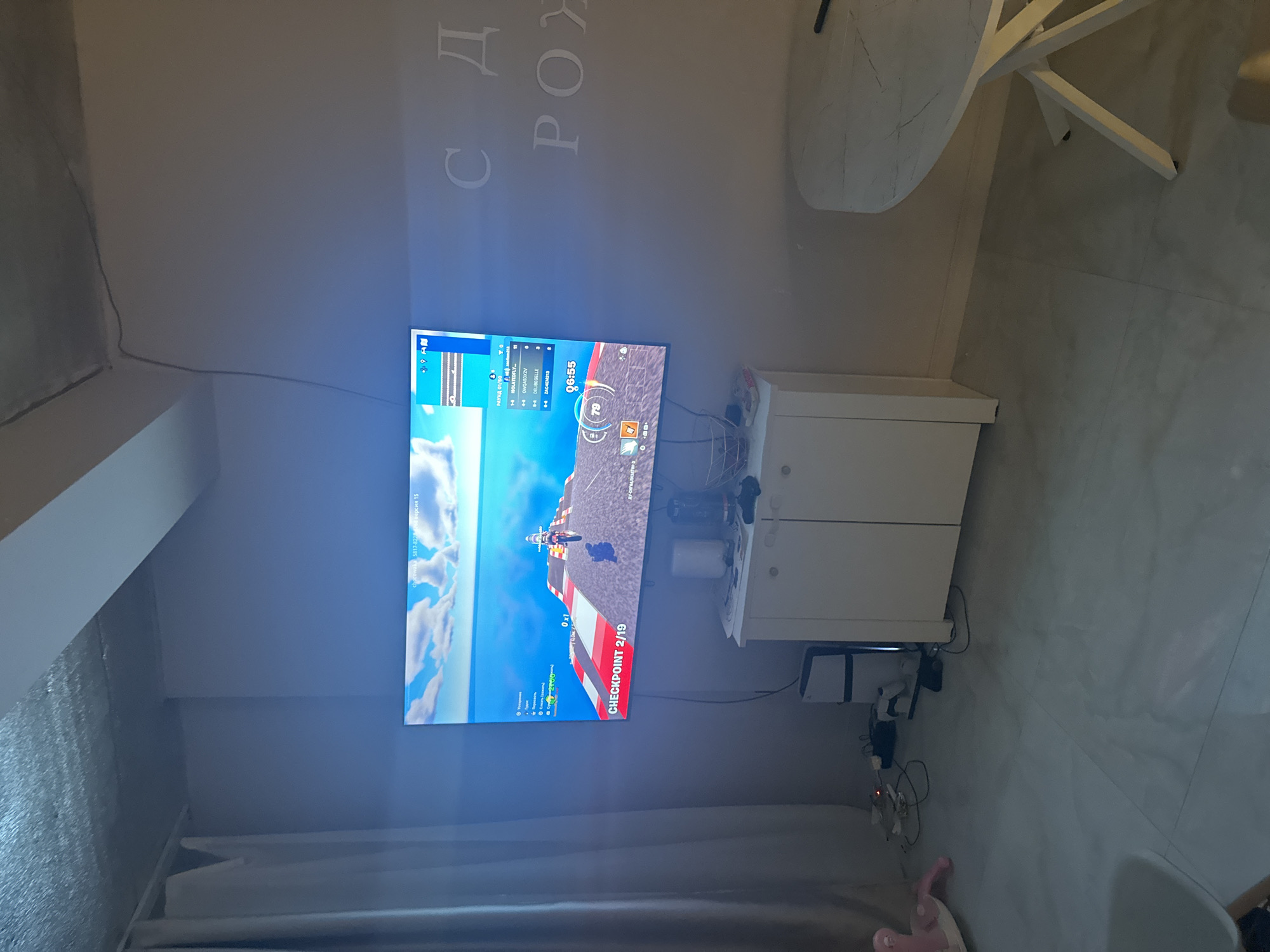 This image captures an indoor scene, likely a living room or bedroom, featuring a large flat-screen television mounted on a wall. The walls are a plain, light color, and the floor is tiled with light-toned rectangular tiles.
The television displays a video game, which appears to be a racing game with a motorcycle visible on a track. Key elements of the game interface include "CHECKPOINT 2/19" at the bottom center, a timer indicating "06:55" in the top right, and what seems to be a player score or level "79". The top left of the screen shows game statistics or player names such as "РАУНД 01/50", "ISOLATEDFLY", "OWQASDXZV", "DELIBEGELLE", and "ZAC4EM213". A game ID or version "ка 5817-8228-4768 версия 15" is also visible.
Below the TV is a white, two-door cabinet. To its right, a white PlayStation 5 console is visible, surrounded by various cables and wires on the floor. Part of a white table and chair can be seen in the upper right portion of the image. Along the bottom of the wall, light-colored curtains hang, and a pink soft toy rests on the floor nearby.
On the wall above the TV, decorative letters in Cyrillic script are partially visible, reading "С Д РОУ". This likely forms part of a phrase such as "С Днём Рождения" (Happy Birthday), providing a cultural cue consistent with the location context of Khimki, Russia. The overall scene suggests a casual home environment where someone is engaged in gaming.