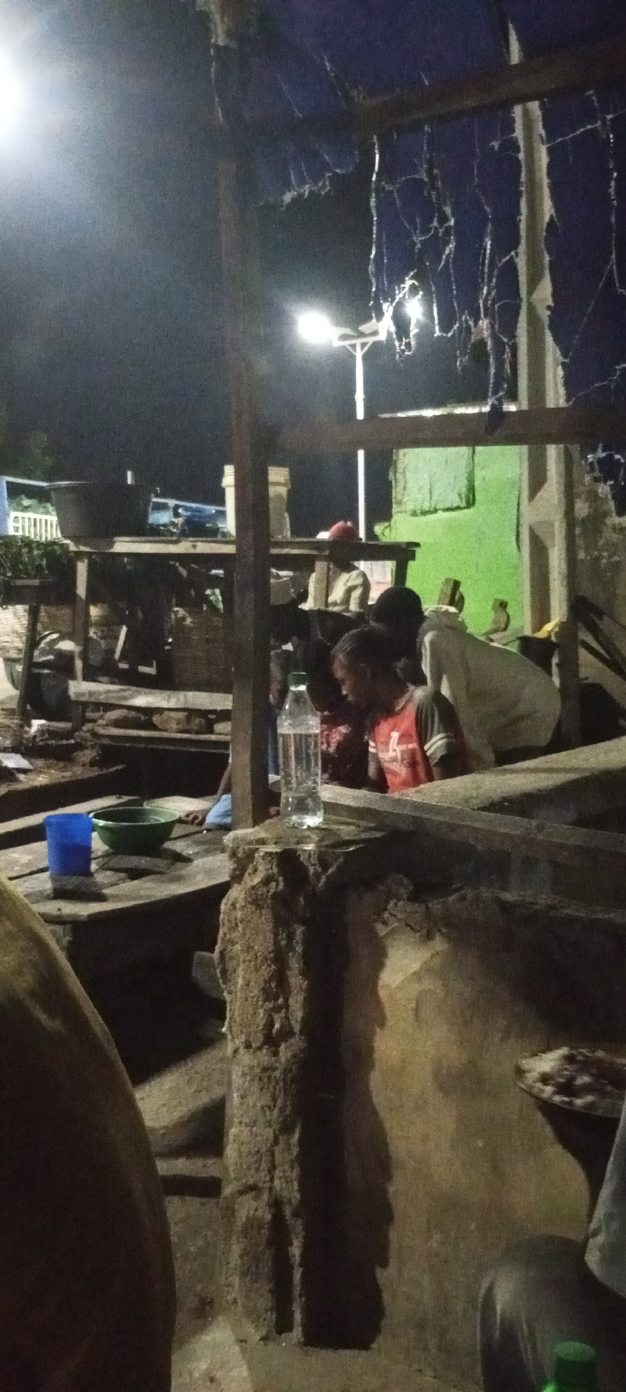 In Adankolo, Nigeria, a dimly lit outdoor scene features a group of individuals gathered around a makeshift wooden structure, possibly a food stall or marketplace, at night. A person wearing a red shirt and a young boy with dark hair are visible in the foreground, seemingly interacting. A partially consumed bottle of water stands on a rough, textured surface. In the background, other figures are present, one wearing a red cap. The environment appears rustic, with evidence of a torn blue tarp or roofing material overhead and a green painted wall in the distance. Wooden tables and baskets are also discernible, suggesting a place of commerce or communal gathering. The lighting is artificial, cast by overhead lamps, creating strong shadows and a somewhat grainy quality to the image.