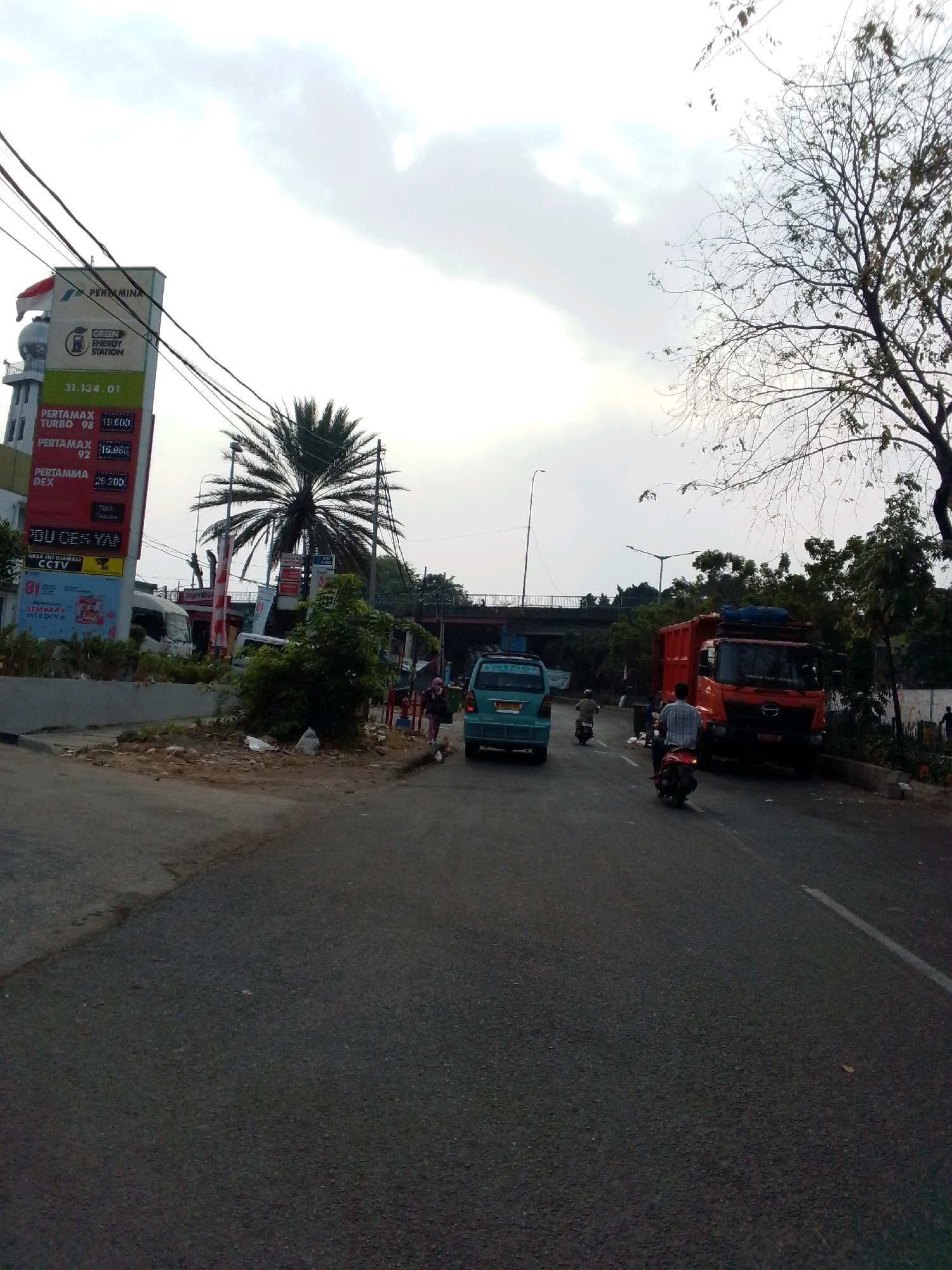 This is a street scene in East Jakarta, Indonesia, with a gas station on the left and a road with traffic in the foreground and middle ground. The gas station has a tall pylon with a Pertamina logo and fuel price displays. There are palm trees and other vegetation around the gas station. The road is asphalt and appears to be a multi-lane road. Traffic includes several cars and motorcycles, as well as a large orange truck. There are people on motorcycles and walking on the sidewalk. The sky is overcast with some sunlight breaking through. The image is taken from a low angle, looking slightly upwards.