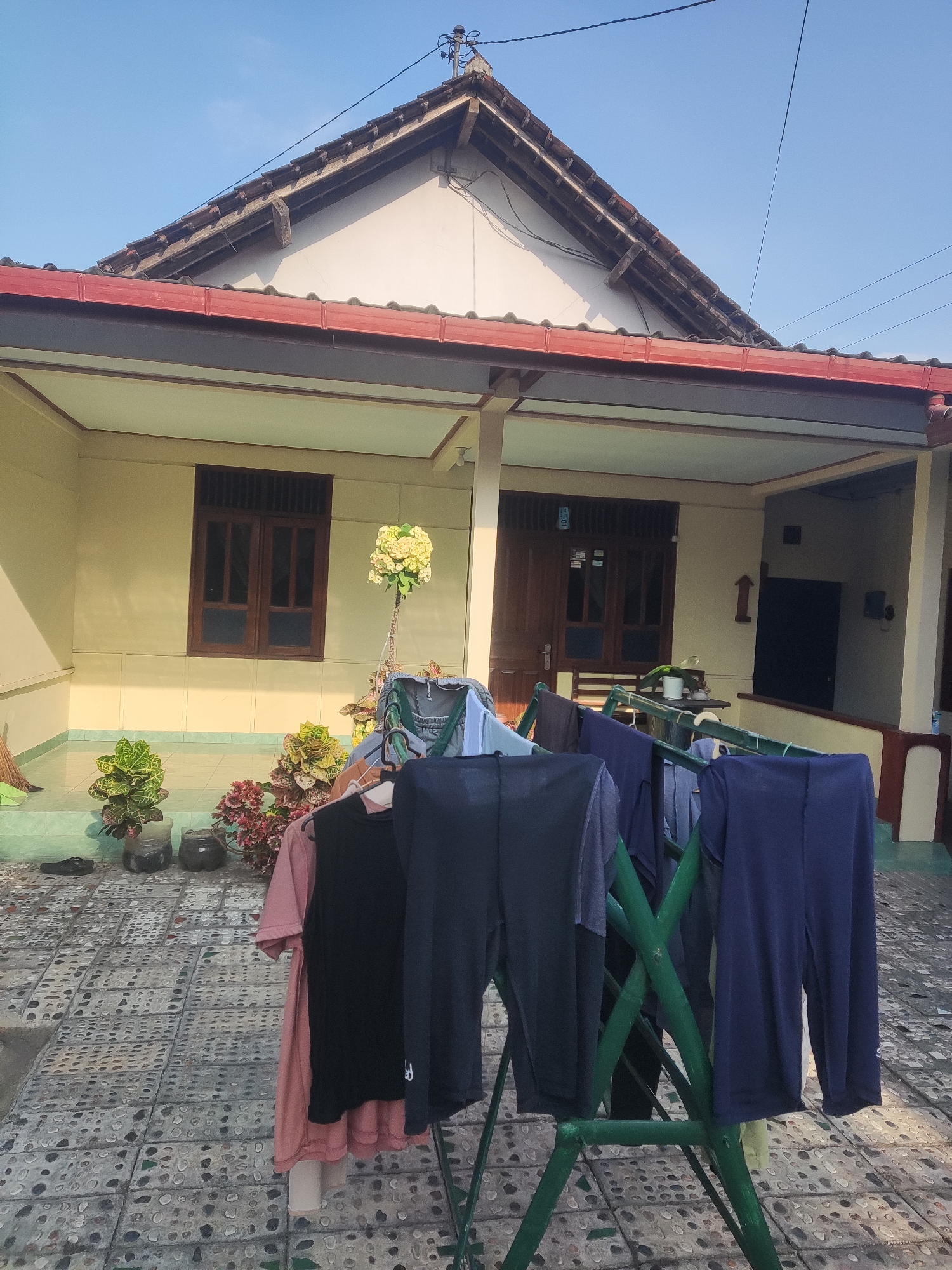 In Trihanggo, Indonesia, a residential scene unfolds under a clear blue sky. A single-story house with a red-trimmed gabled roof and pale yellow siding dominates the background. Dark brown wooden windows with grilles are visible on the facade, along with a wooden front door. In the foreground, a green clothes drying rack holds an assortment of dark-colored clothing, including pants and shirts. The ground is paved with a patterned tile mosaic. Several potted plants, including a flowering ornamental plant, are arranged near the house. A broom leans against the wall on the left side of the image. The overall impression is one of domestic life in a tropical setting.