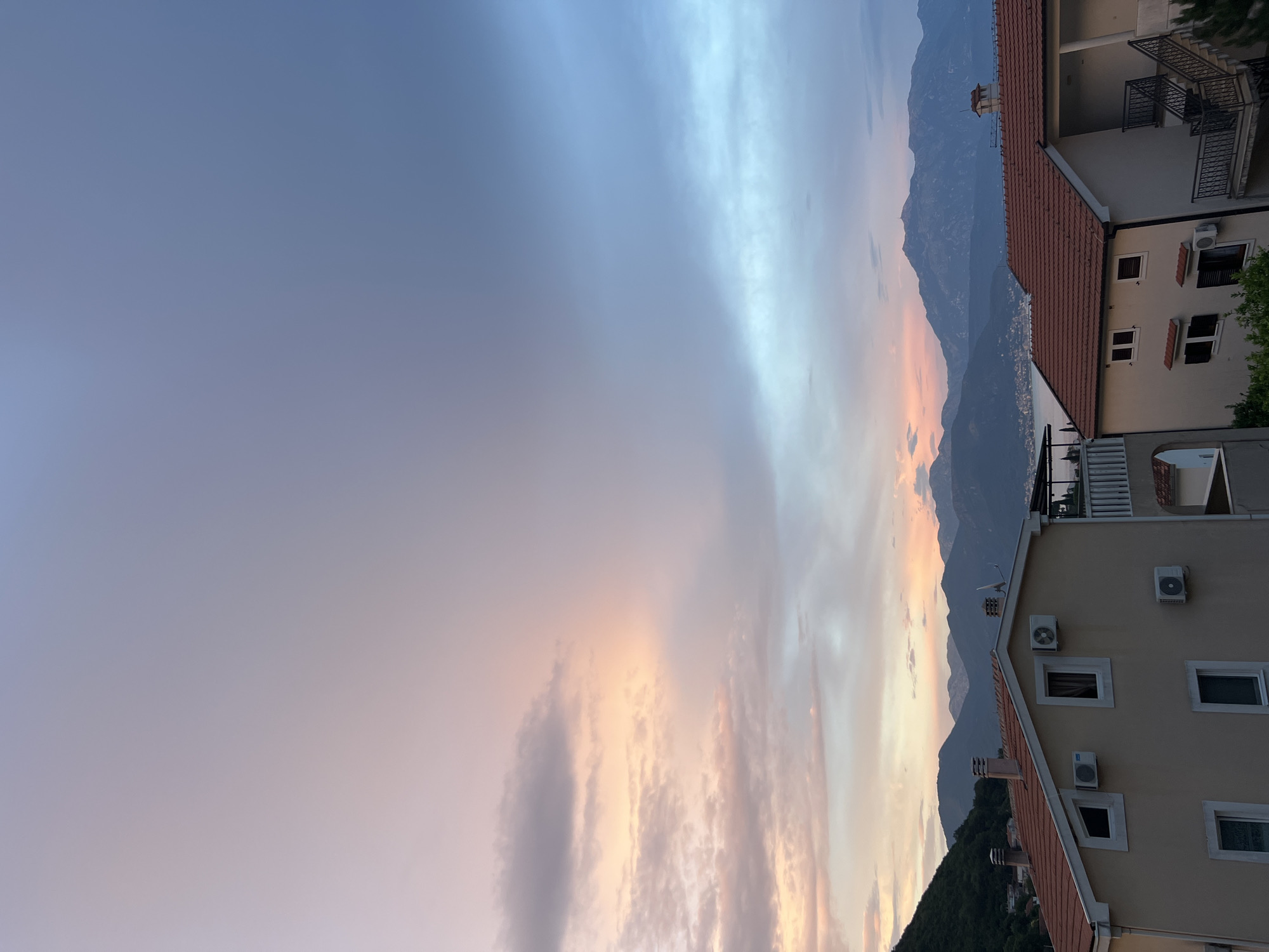 The image shows a picturesque sunset over a coastal town in Baošići, Montenegro. The sky is painted with soft hues of blue, pink, and orange, with wispy clouds stretching across the horizon. In the foreground, a multi-story building with a tiled roof is partially visible, featuring several windows and air conditioning units. To the left, lush green hills descend towards the sea, which is faintly visible in the distance. Towering mountains form a dramatic backdrop, their silhouettes softened by the fading light. The scene evokes a sense of tranquility and natural beauty, characteristic of a peaceful evening by the sea. The time of day is clearly sunset, indicated by the warm, diffused light and the colors in the sky. No people or text are visible in the image.