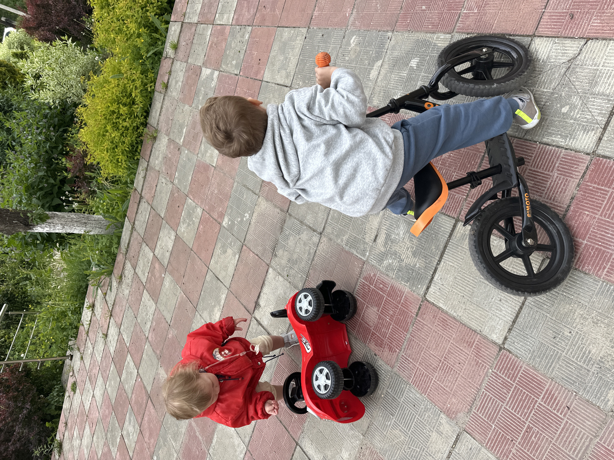 Two children are on a paved area, likely a sidewalk or backyard, constructed of alternating red and gray square tiles. The child on the right, wearing a gray hooded sweatshirt and blue pants, is riding a black balance bike with orange accents. The child on the left, wearing a red jacket, is playing with a red plastic toy car. Lush green bushes and foliage are visible in the background, suggesting a garden or park setting. There is no way to verify if the image location is Nizhny Novgorod, Russia.